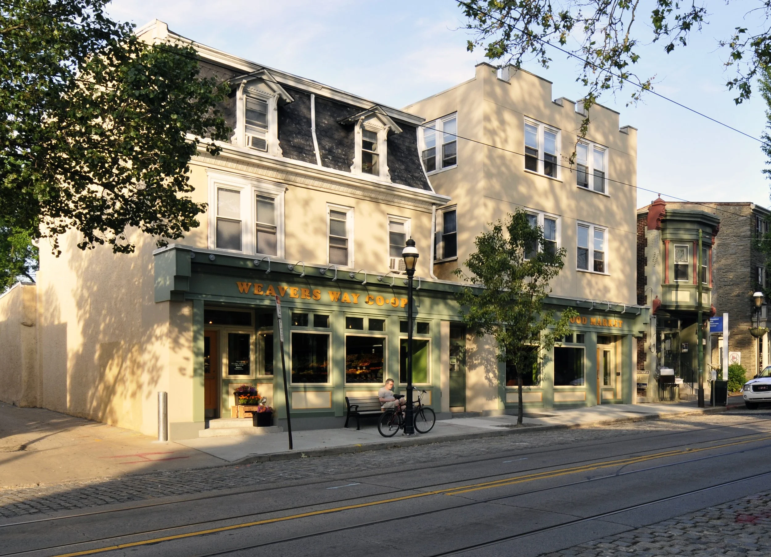 Street view of a building with a storefront named Weavers Way Co-Op, trees lining the sidewalk, a bench with a person sitting, and a bicycle parked near a lamppost.