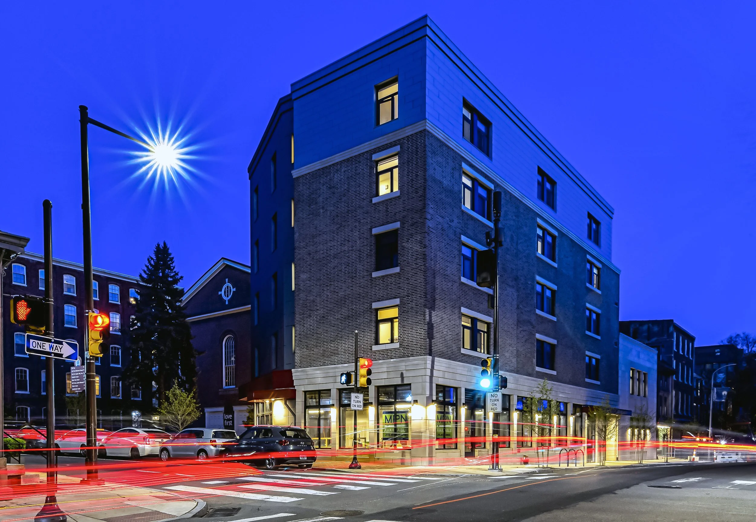 A city street scene at night with a modern multi-story brick building, traffic lights, moving cars creating light streaks, traffic signs, parked cars, and illuminated windows against a dark sky, with a bright full moon.