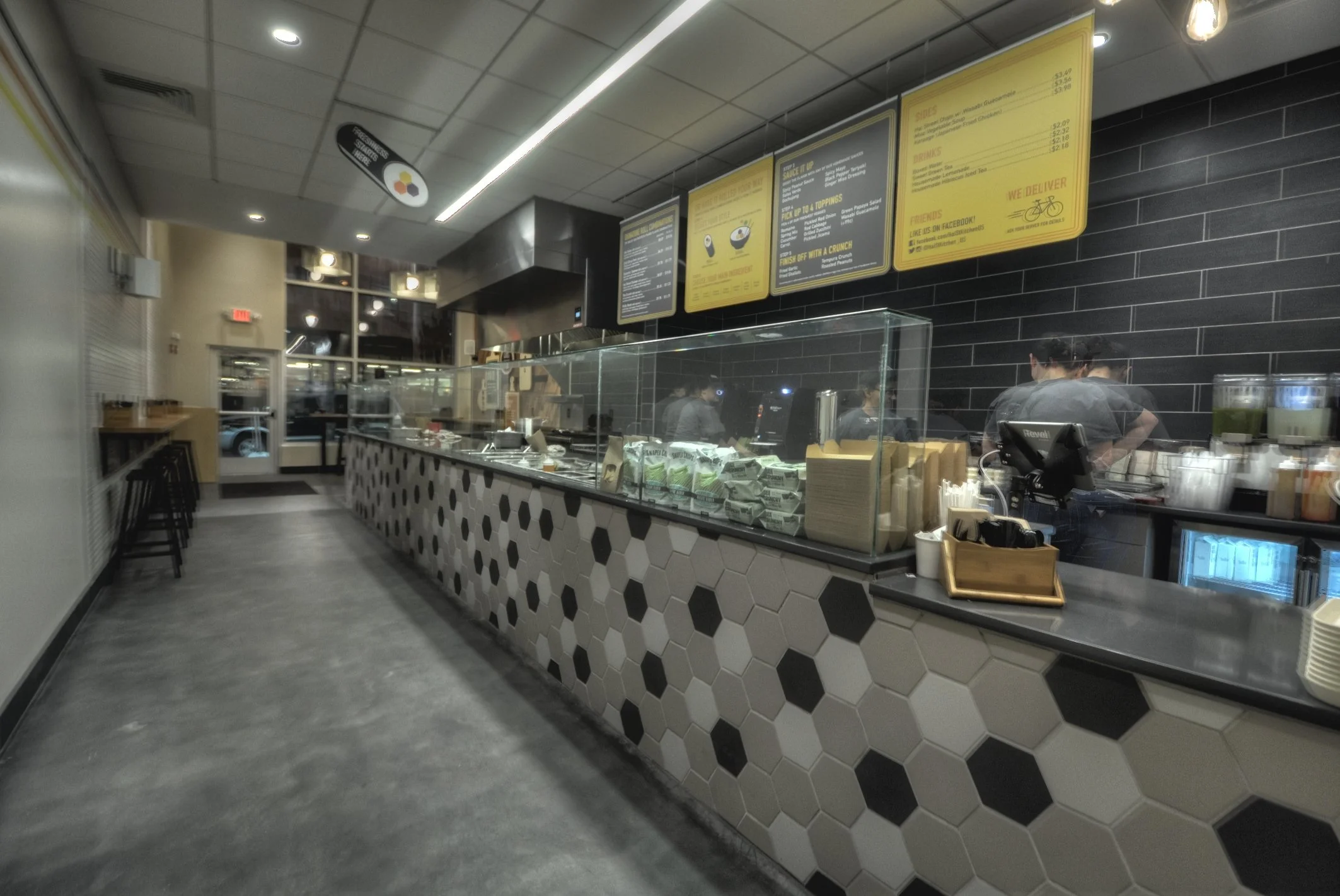 Interior of a modern fast food restaurant with black and white hexagonal tile design on the counter, digital menu boards above, and a visible kitchen area behind the counter with staff preparing orders.