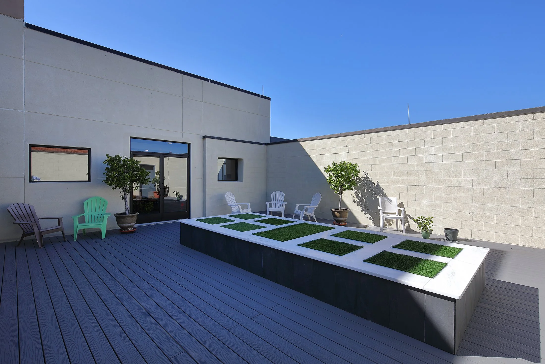 Empty rooftop terrace with wooden decking, small trees in pots, white and purple chairs, and rectangular patches of artificial grass on a raised platform, against a beige building wall under a clear blue sky.