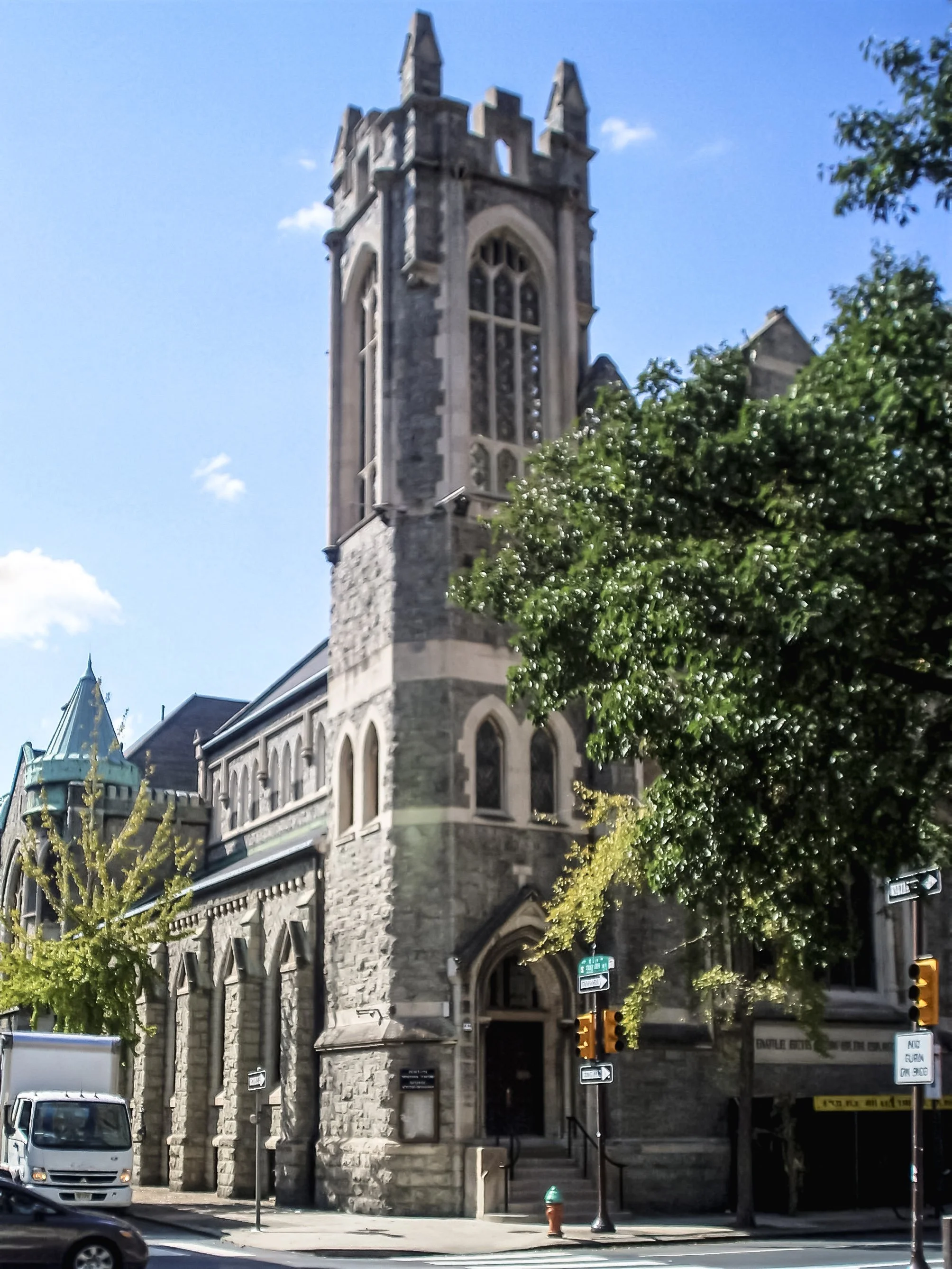 A stone church with a tall, narrow bell tower and Gothic style windows on a city street corner on a sunny day, with a crosswalk, cars, and street signs visible in the foreground.