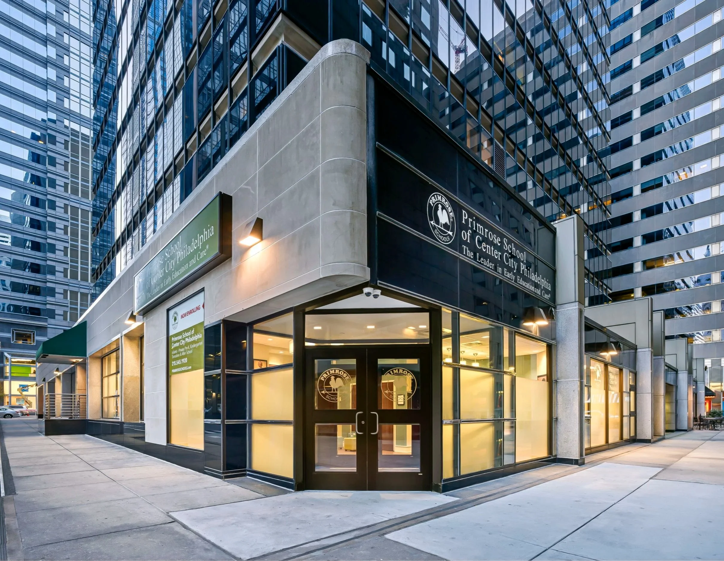 Exterior view of Primrose School of Center City Philadelphia, a modern commercial building with glass windows and a corner entrance, illuminated in the evening.