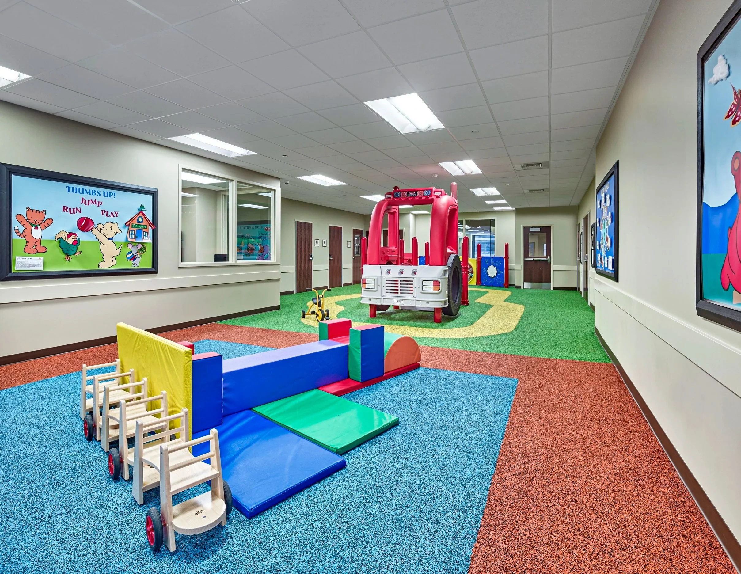 Interior view of a children's play area with colorful padded mats, a small wooden toy cart, a red and white plastic play truck, and wall-mounted screens displaying cartoon characters.