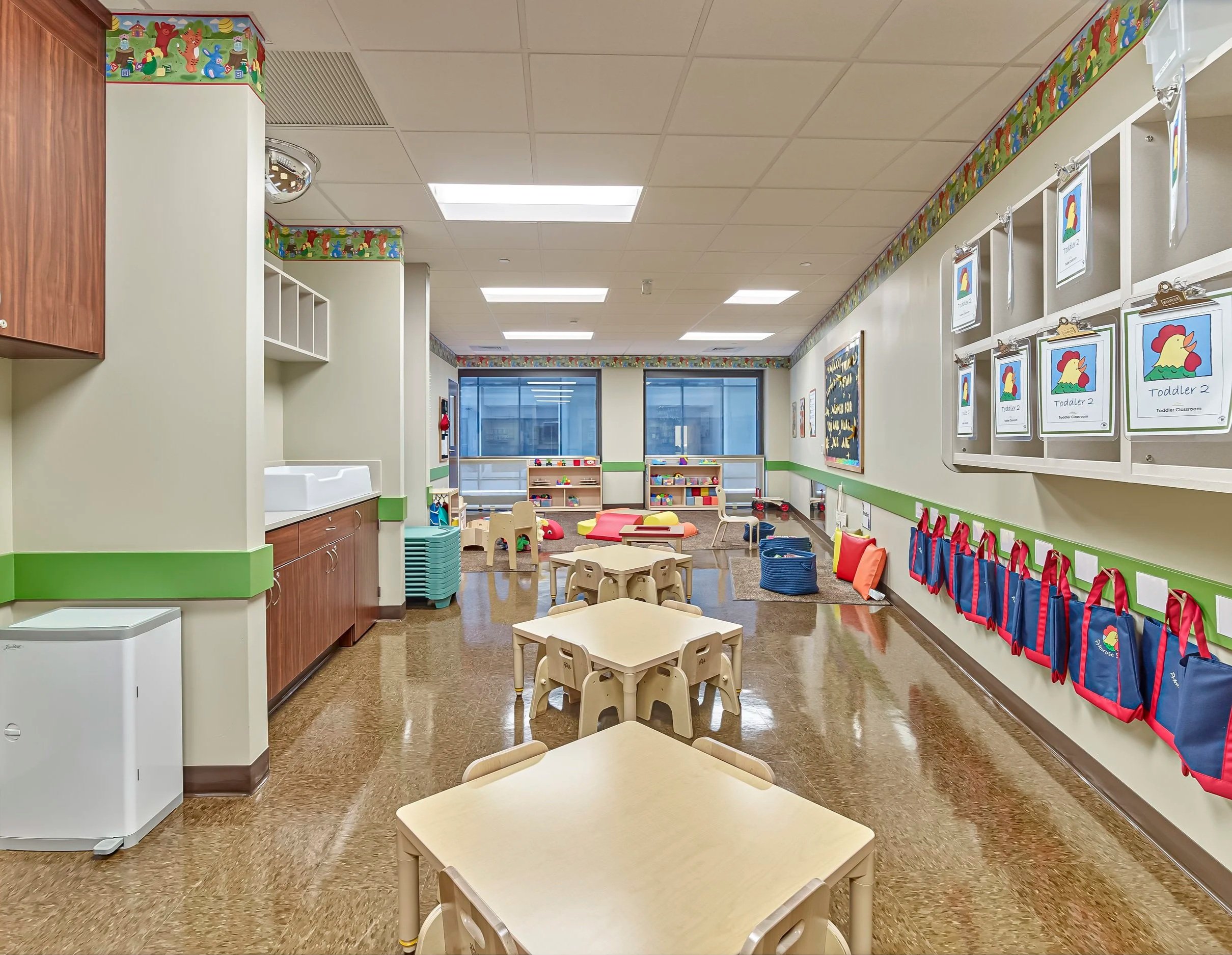Empty classroom in a preschool or daycare with small tables and chairs, colorful storage bins, and educational posters on the walls.