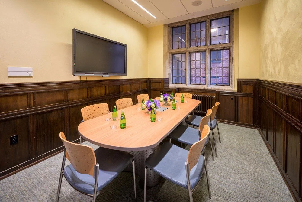 Meeting room with a long wooden table, eight chairs, a large window, flowers, green bottles, and a wall-mounted TV.