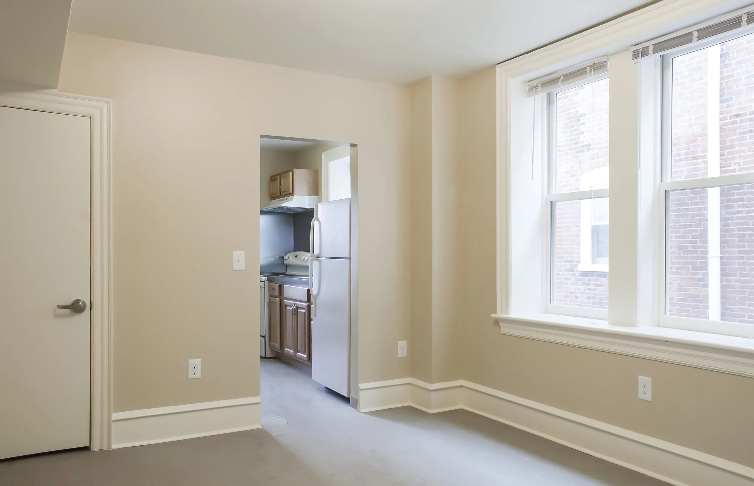 Empty living room with beige walls, a large window with white blinds, and an open doorway leading to a kitchen with wooden cabinets and white appliances.