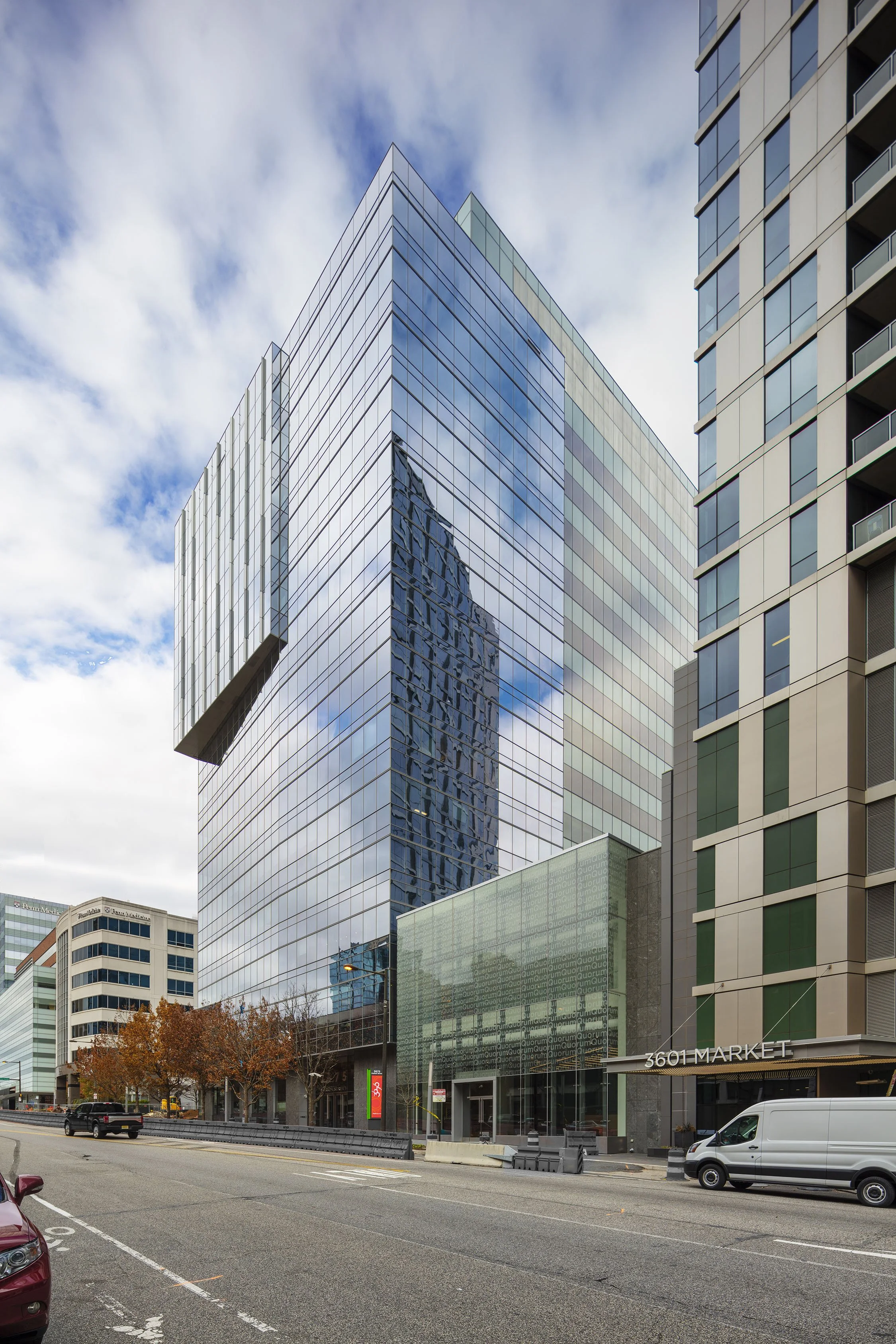 Modern glass office building reflecting nearby buildings and sky, located at 3601 Market Street, with a street view including cars and trees with autumn foliage.