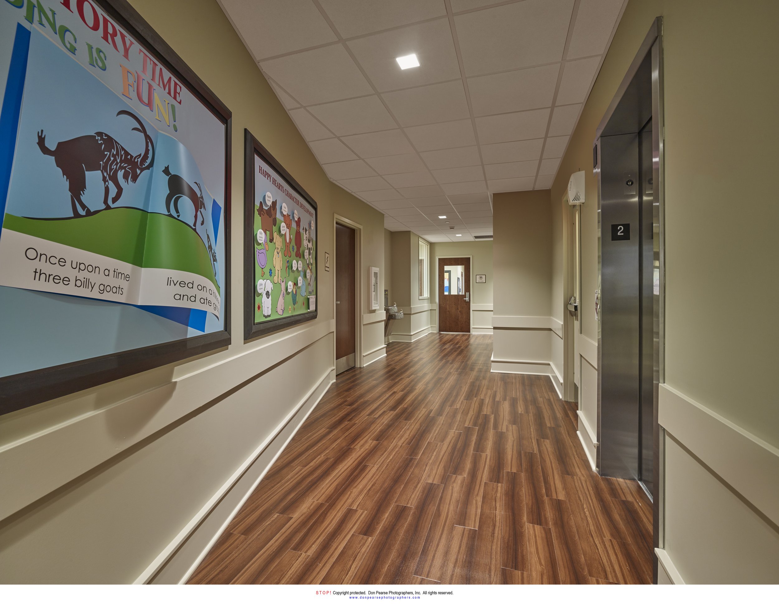A hallway with wooden flooring, decorated with children's artwork and posters, including a storytime theme and a Christmas display. An elevator with the number 2 is on the right side, and natural light is coming from the windows at the end of the hal
