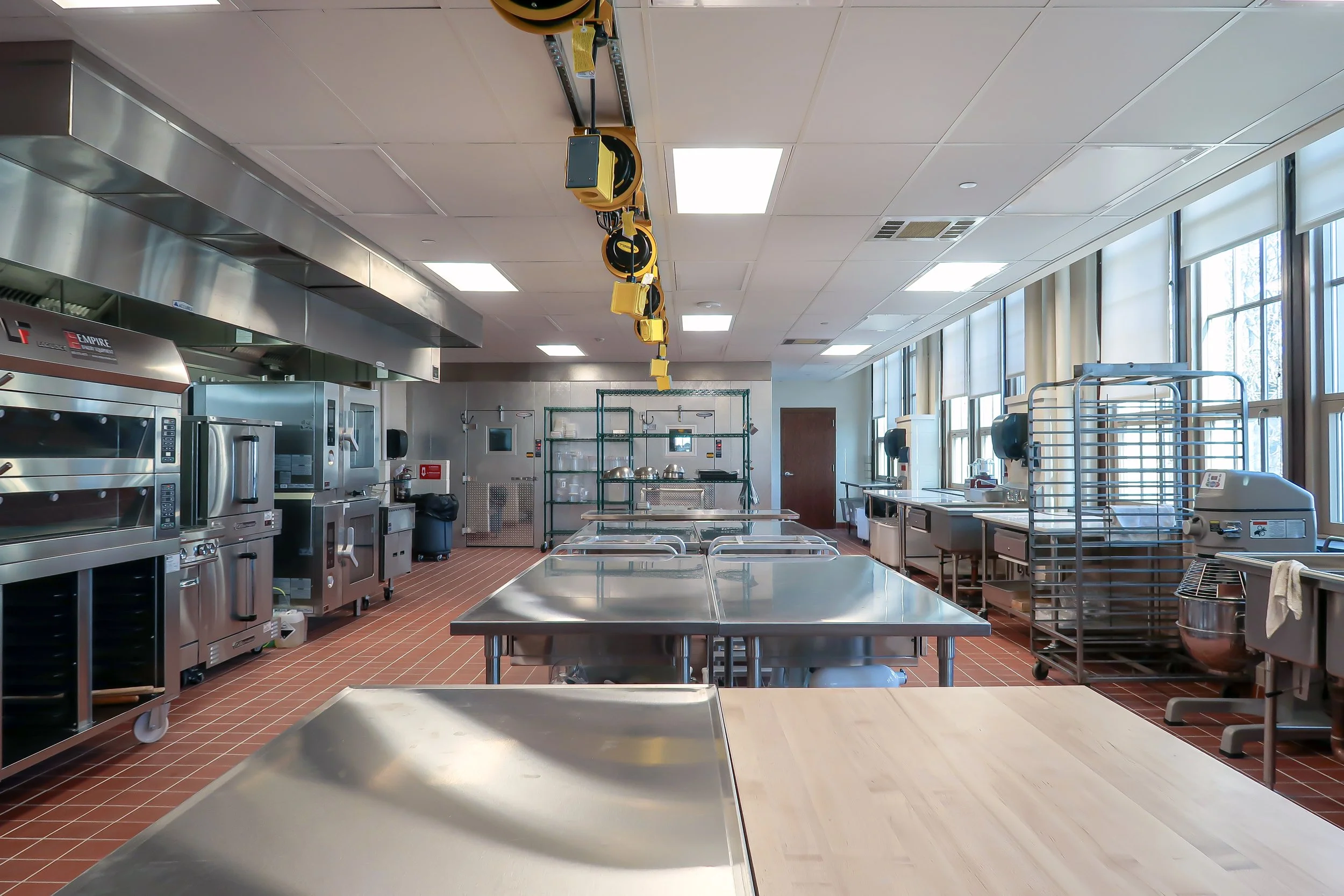 Empty commercial kitchen with stainless steel appliances, worktables, stainless steel shelves, baking racks, and large windows along one side.