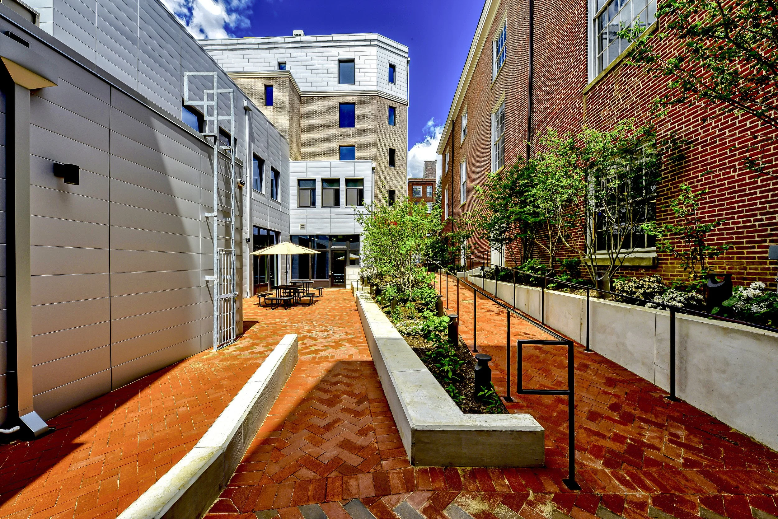 An outdoor courtyard area between modern and brick buildings with trees, garden beds, a picnic table with an umbrella, brick pavement, and metal railing.