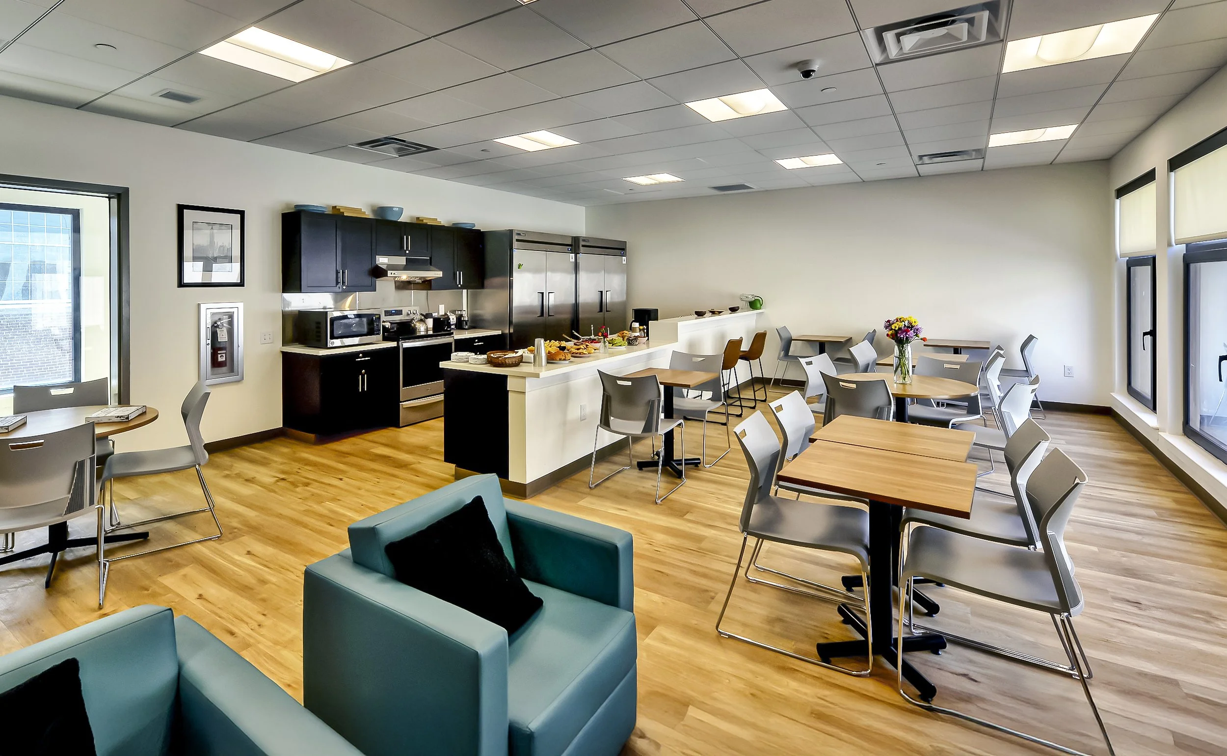 A modern communal kitchen and dining area with wooden floors, black cabinets, stainless steel appliances, a breakfast bar, and tables with chairs, decorated with a vase of flowers near large windows.