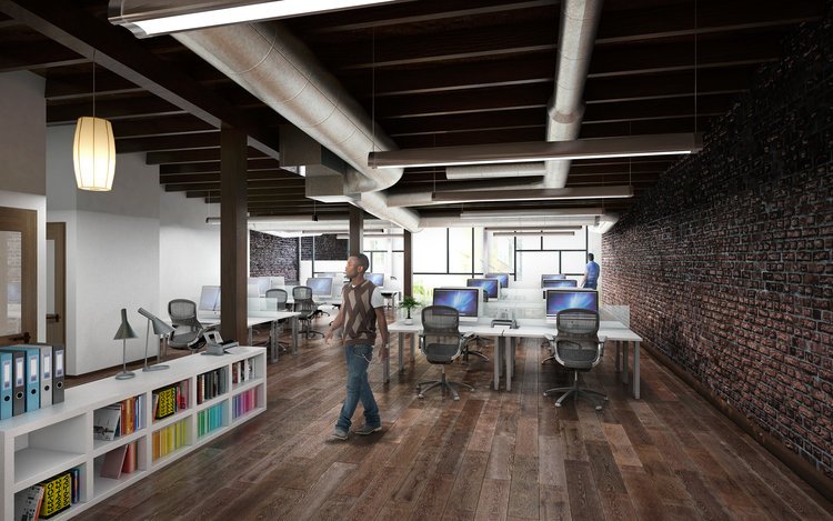 Modern open office with exposed brick wall, wooden floors, and ceiling pipes, featuring white work desks with computers, black office chairs, a bookshelf with colorful binders, and a person walking through the space.