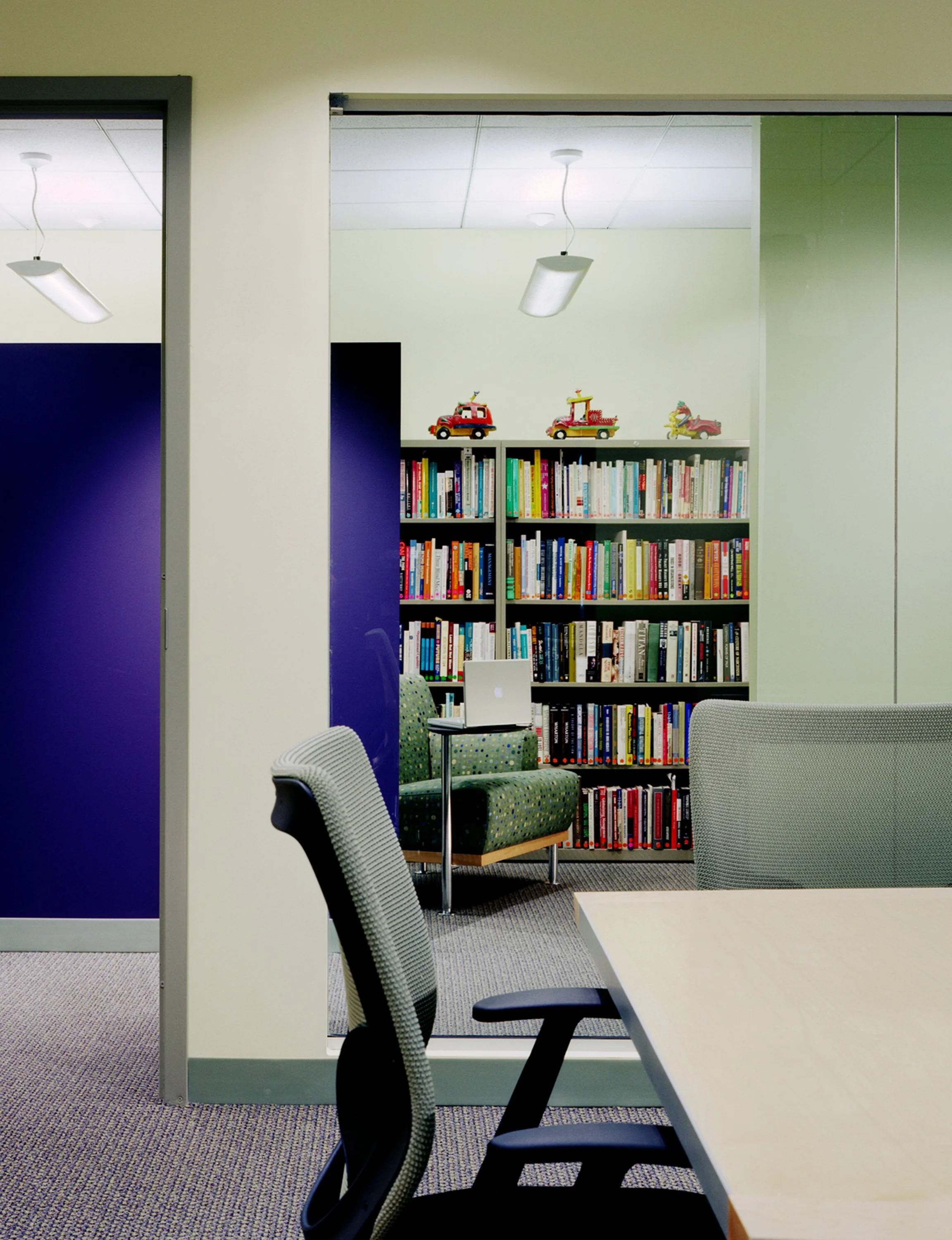 View of a room with a seating area containing a green armchair with a small table holding a laptop, and a bookshelf with colorful books and toy vehicles on top, seen through a glass wall.