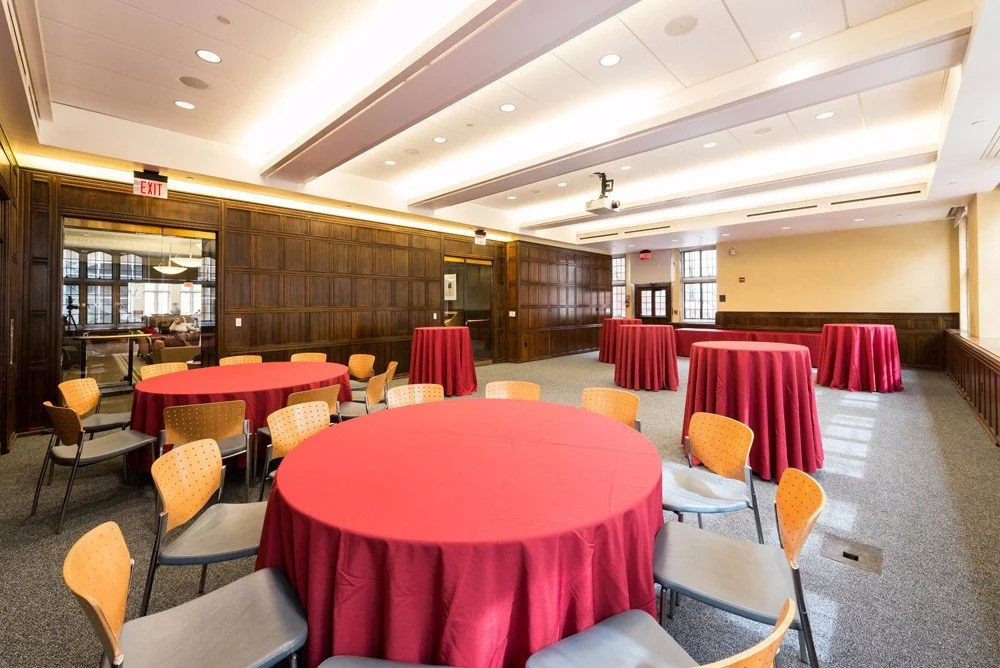 Event room with round tables covered in red tablecloths, surrounded by yellow and gray chairs, with wood-paneled walls and large windows sending natural light into the space.