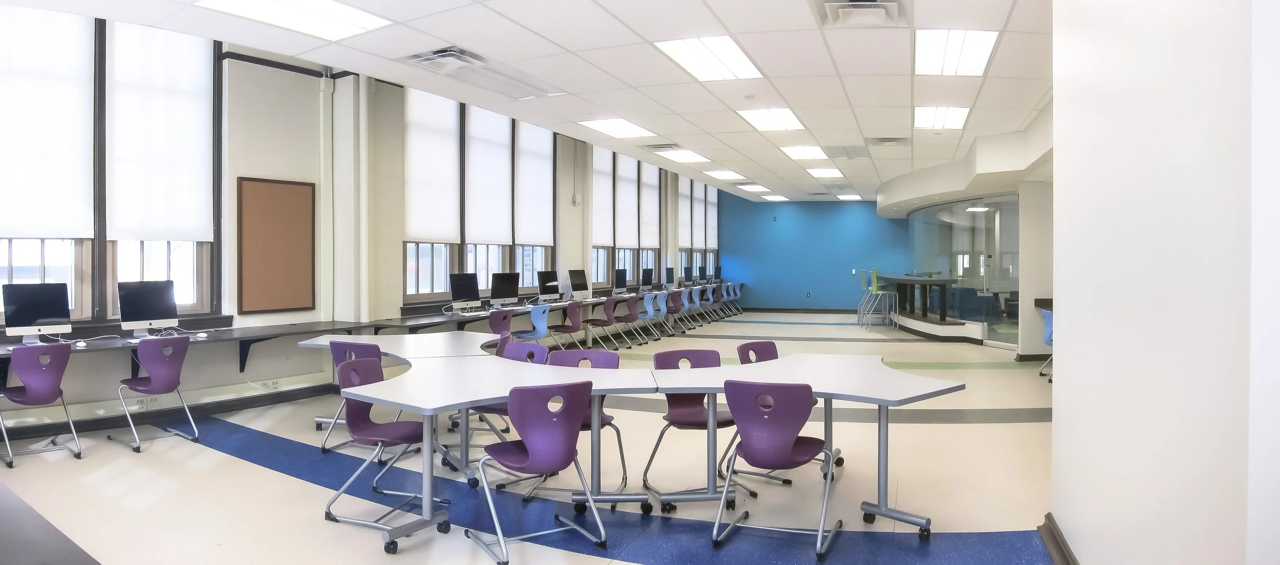 Empty classroom with large windows, purple chairs, and computers along the wall. There is a blue accent wall and a tiered seating area with green chairs.