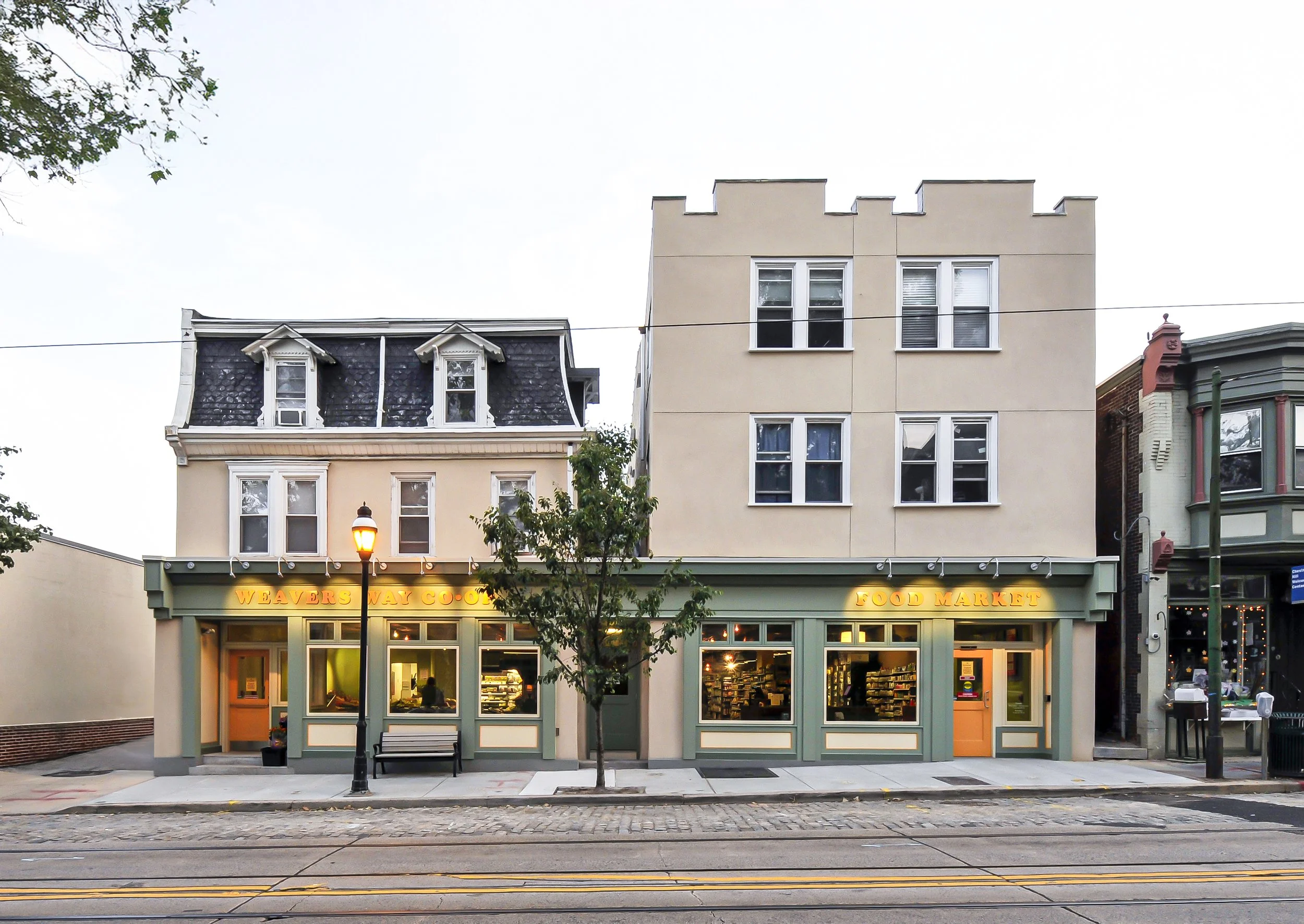 Street view of a small retail building with a green facade, displaying the signs 'Weavers Way Co-op' and 'Food Market'. The building has three large windows with interior lighting, a door on the right, a bench, and a streetlamp in front. Surrounding 