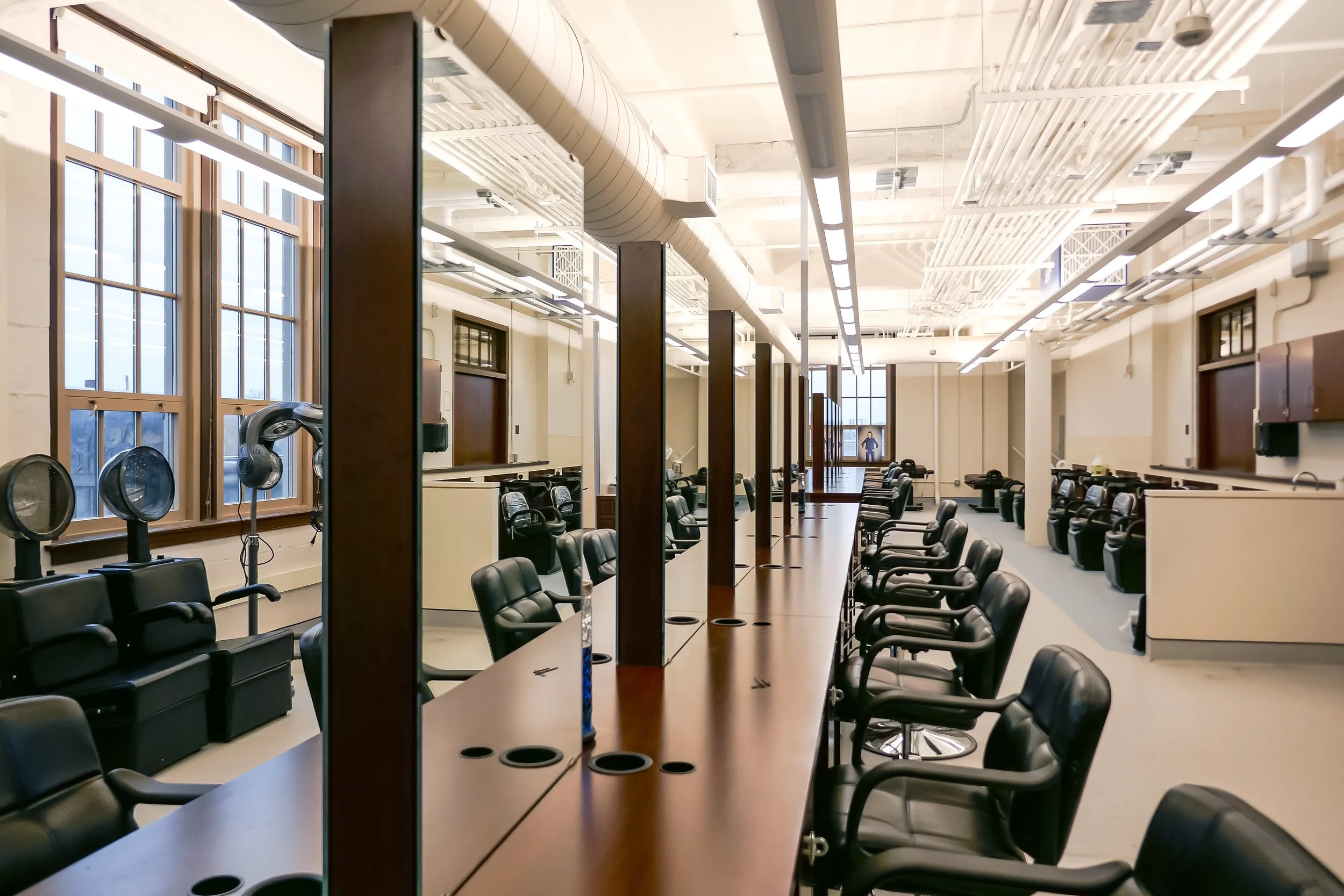 A large, modern hair salon with multiple styling stations, black chairs, large windows, and industrial ceiling design.