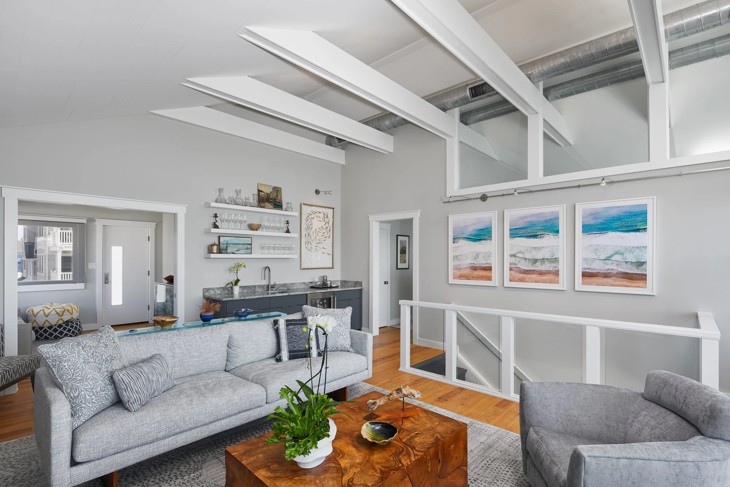 Living room with gray couch, armchair, wooden coffee table, artwork of ocean waves on wall, open kitchen, and white walls with ceiling beams.