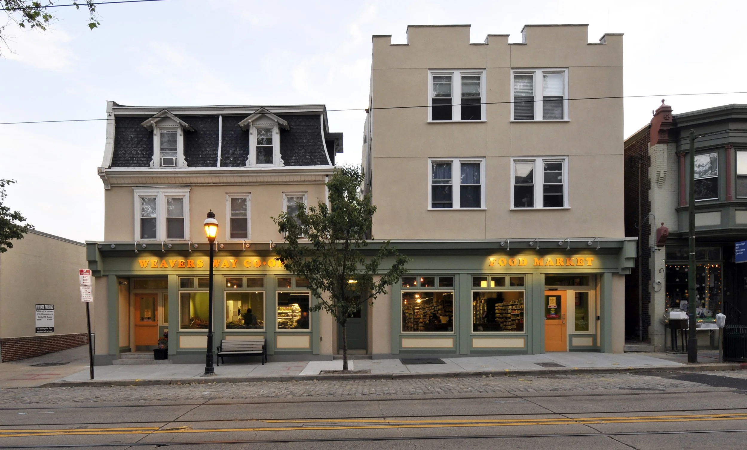 Street view of a small retail building with a green facade, displaying the signs 'Weavers Way Co-op' and 'Food Market'. The building has three large windows with interior lighting, a door on the right, a bench, and a streetlamp in front. Surrounding