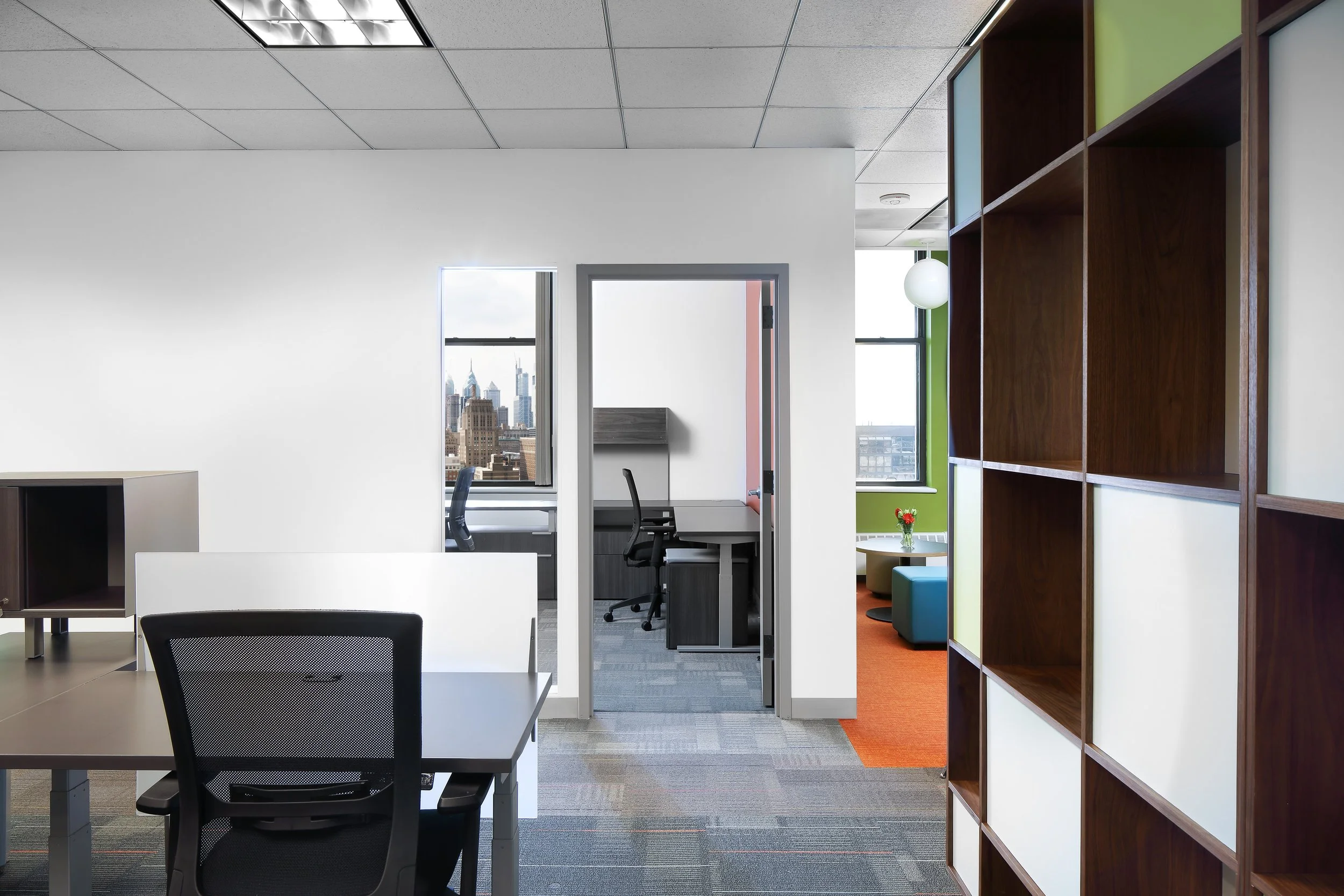 Modern office space with desks, chairs, a bookshelf, and city view windows.