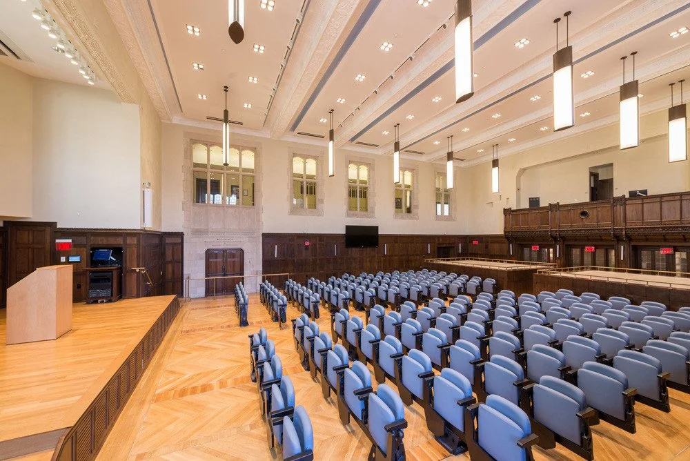 Large conference room with rows of blue chairs facing a stage with a wooden podium, in a well-lit space with high ceilings, chandeliers, and large windows.