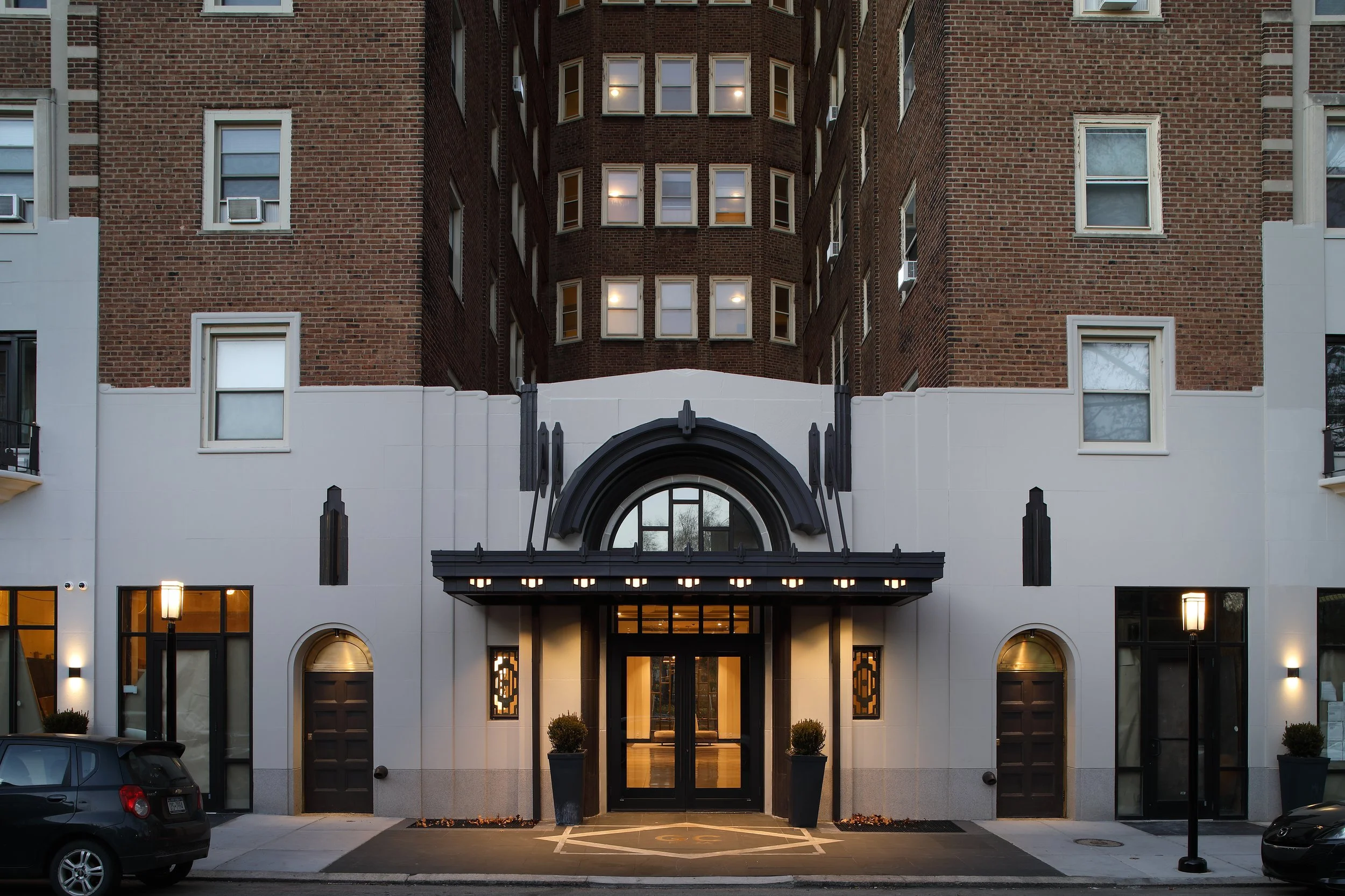Front view of a modern urban building with a black entrance door, large windows, and decorative black accents, with cars parked in front and street lamps illuminating the sidewalk during dusk.