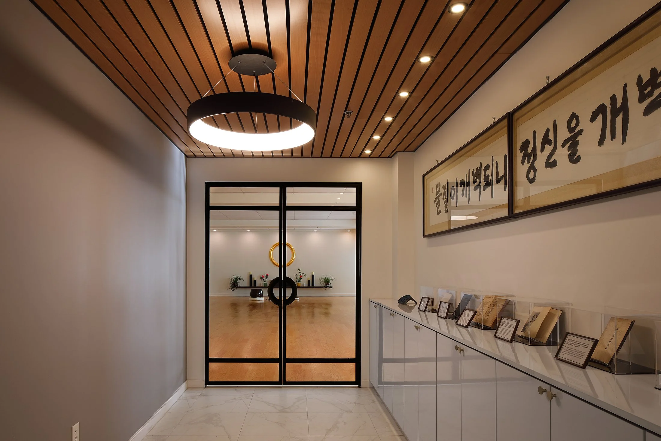 Interior view of a modern building with a hallway leading to a room, featuring wooden ceiling, black framed glass doors, framed Korean calligraphy art on the right wall, and display cases with books and framed information.
