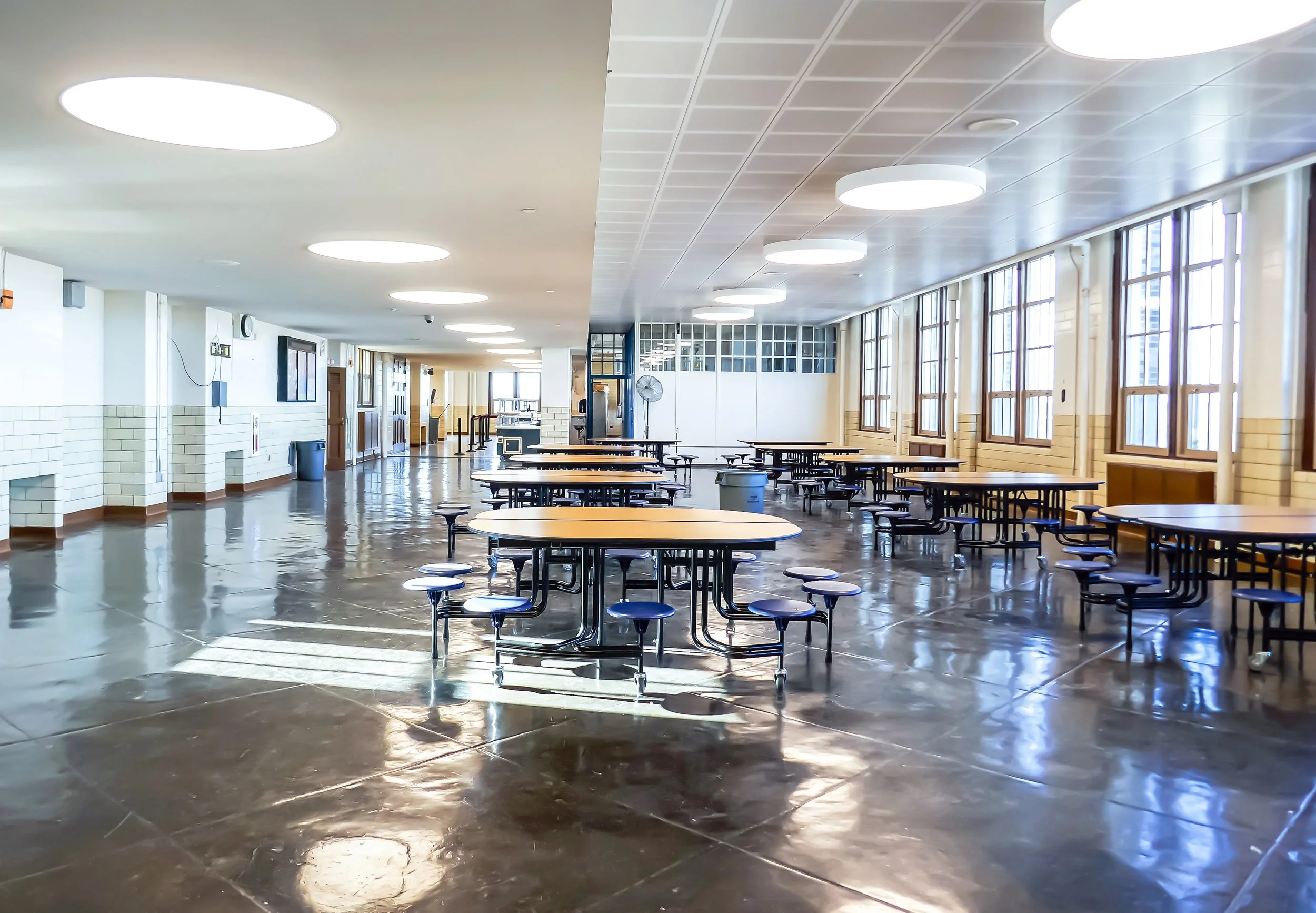 Empty cafeteria with round tables and attached benches, large windows along one wall, ceiling lights, polished floor, and doors at the far end.