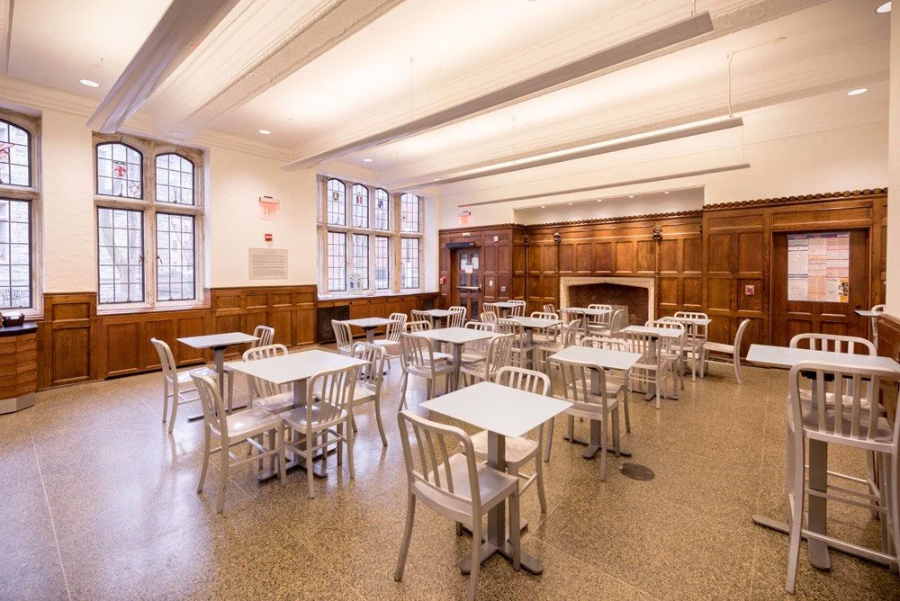 Empty dining room with white tables and chairs, wooden paneling, large stained glass windows, and a fireplace.