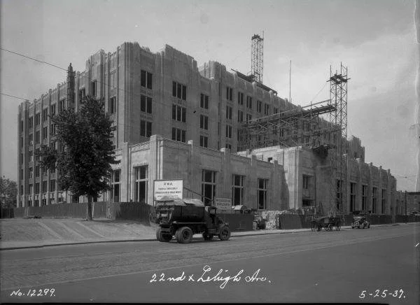 Black and white photo of a large building under construction, with scaffolding and cranes visible. Several early 20th-century cars are parked along the street in front, with a tree on the left side. Handwritten note at the bottom reads "22nd and Lehi