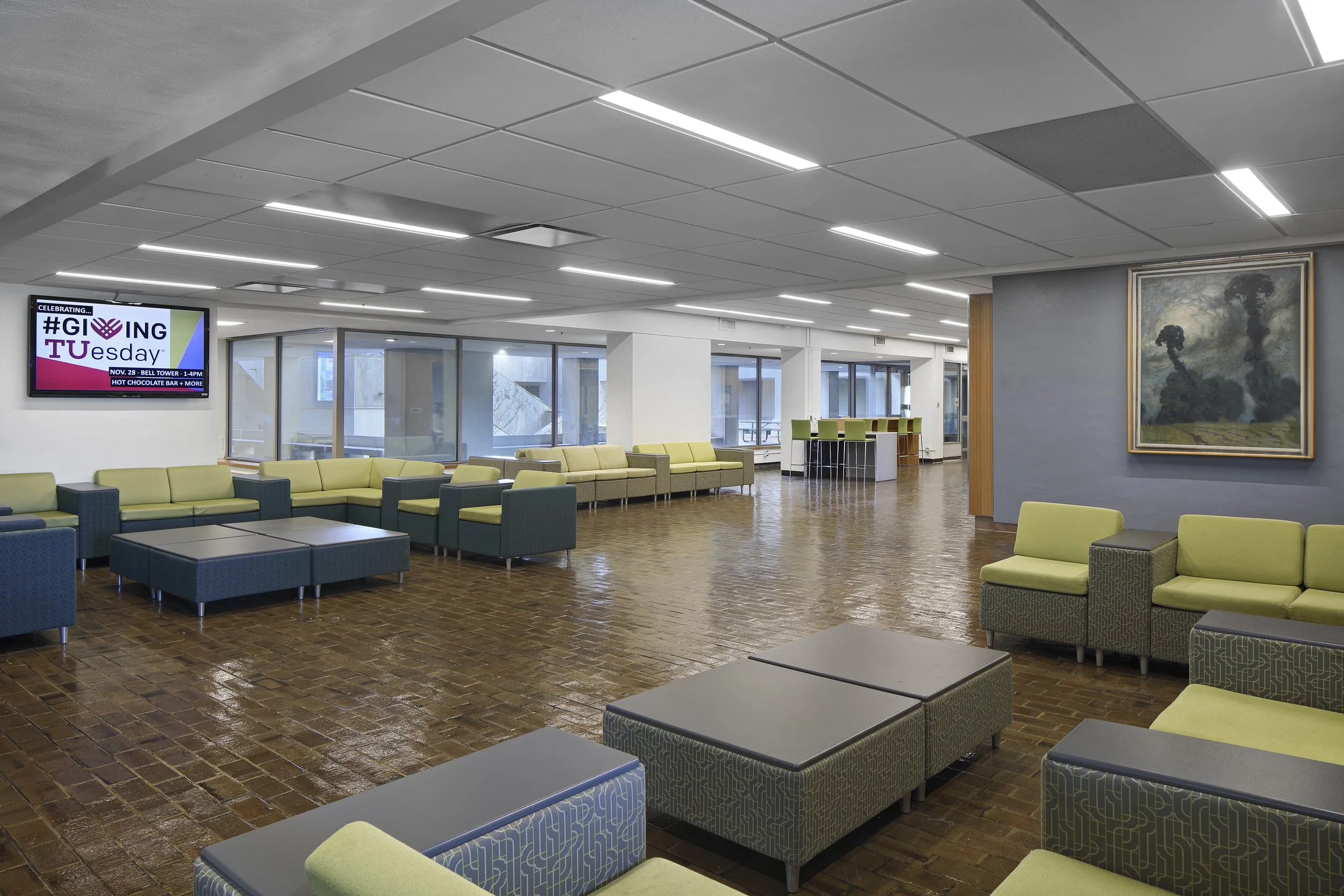 Modern lobby with green and blue seating, wood flooring, large windows, and a flat-screen TV displaying #GivingTuesday event information.