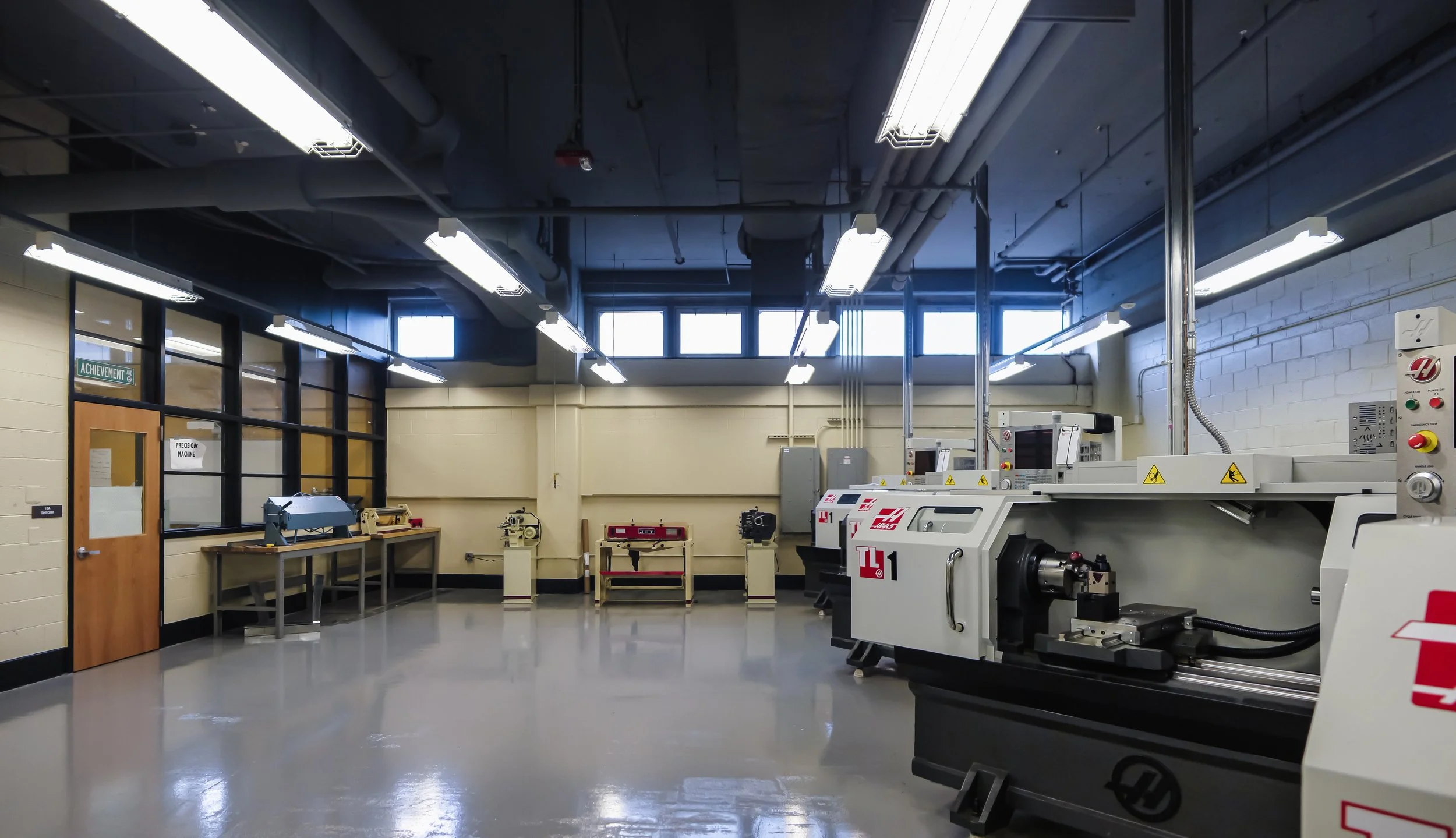 Machine shop with various precision machinery, workbenches, and windowed wall, illuminated by overhead fluorescent lights.