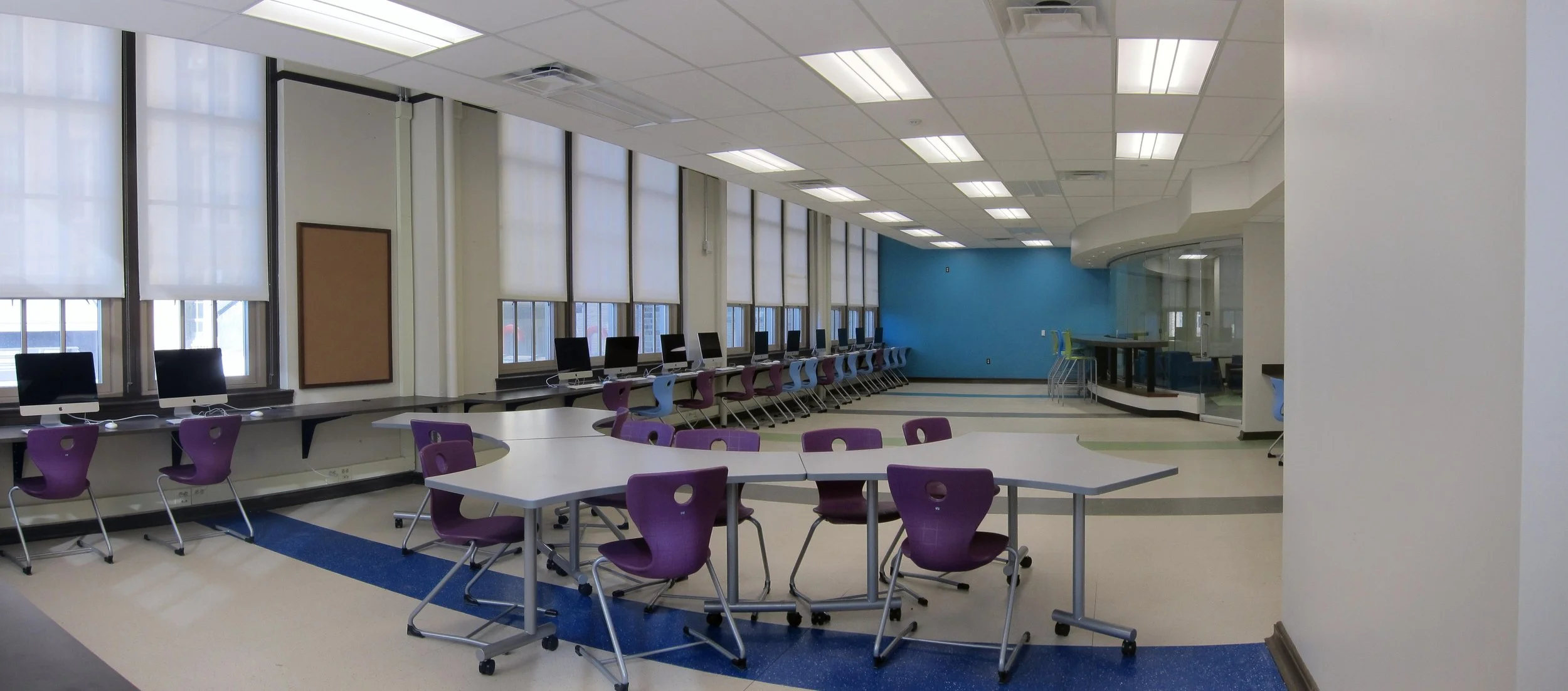 Empty classroom with large windows, purple chairs, and computers along the wall. There is a blue accent wall and a tiered seating area with green chairs.