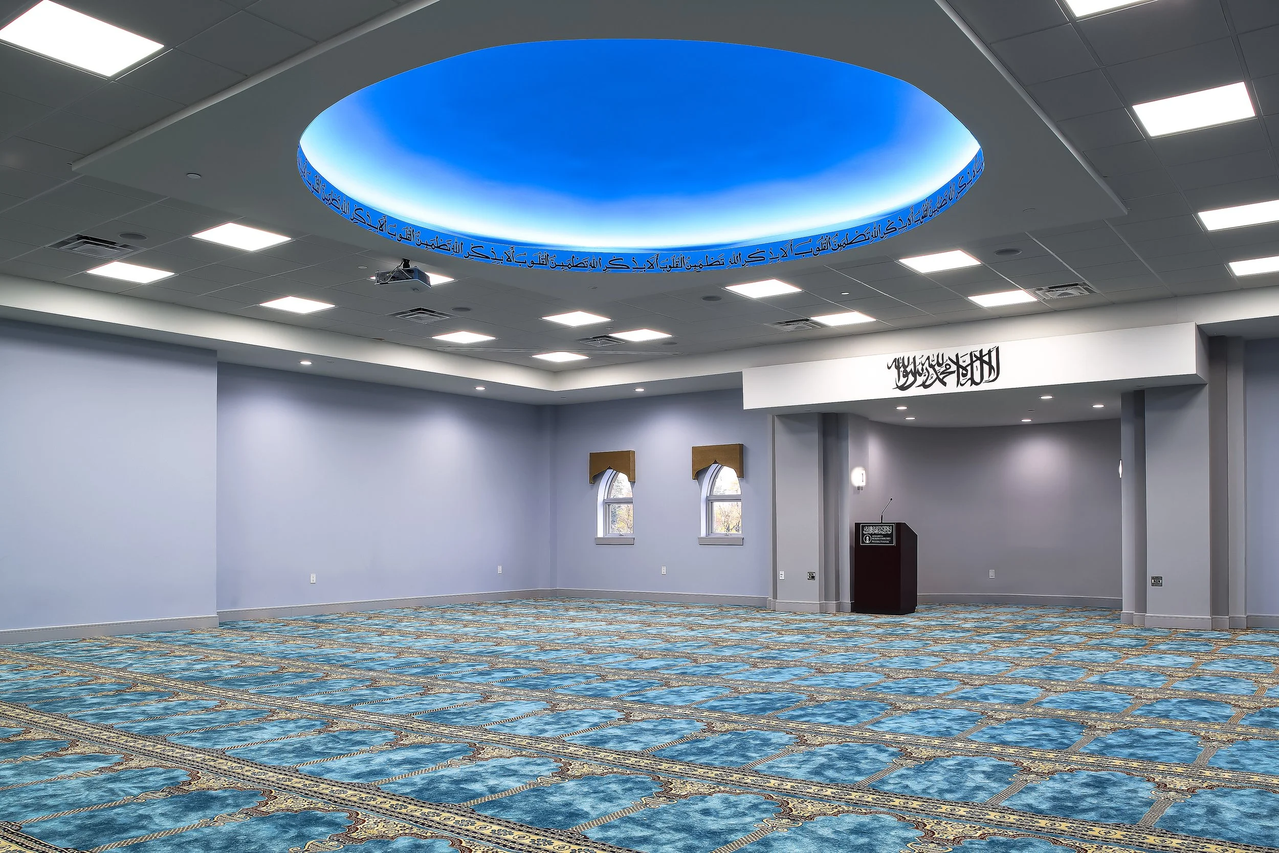 Empty prayer room with blue patterned carpet, light blue walls, two small windows with gold valances, a central blue ceiling dome with Arabic script, and an elevated area with a sign and podium.