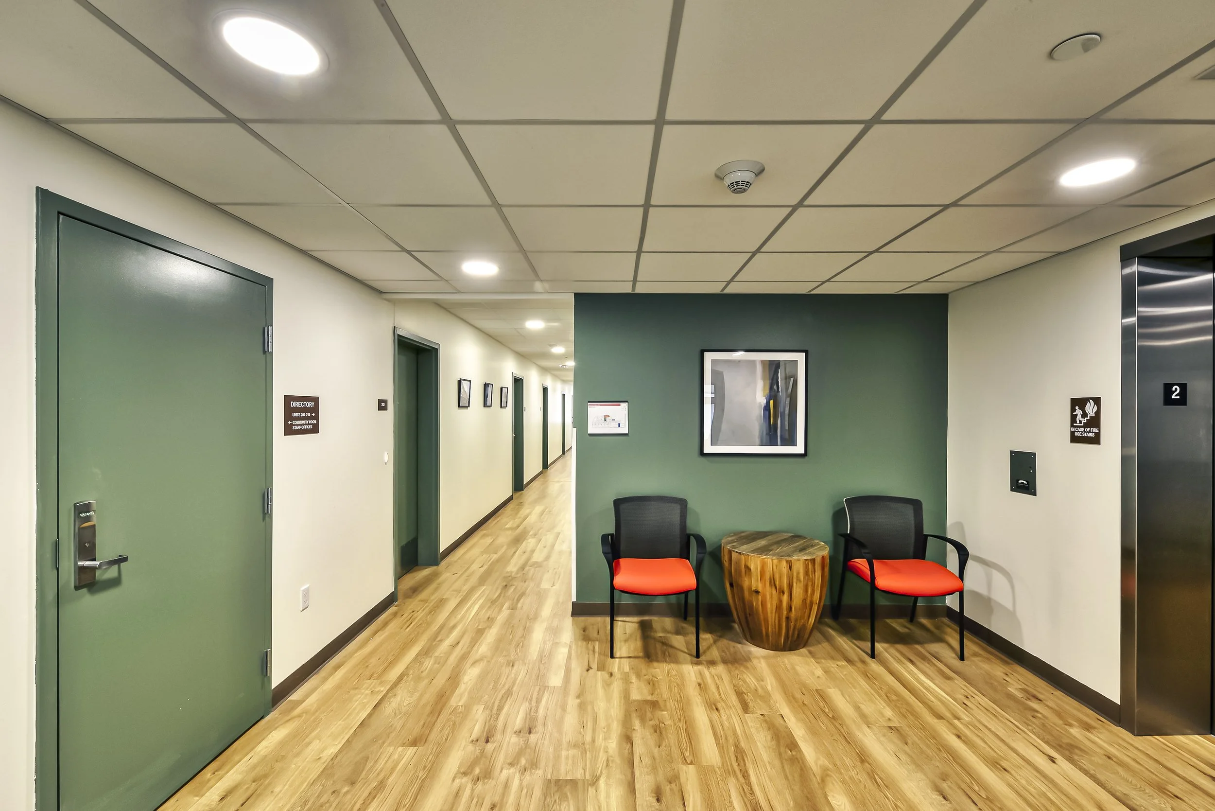 A corridor with doorways and framed pictures, leading to an area with two black chairs with red cushions and a wooden side table, on a green accent wall in a building interior.