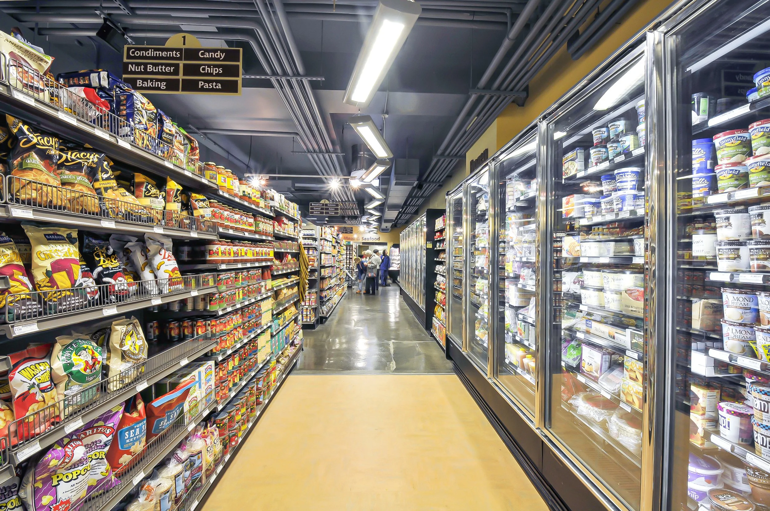 Aisle in a grocery store with shelves stocked with snack foods, condiments, and refrigerated dairy products, with customers shopping in the background.