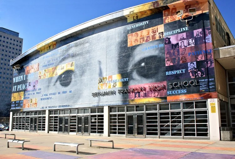Large mural on a building featuring a giant close-up of a person's eyes with various words and images related to education and values, such as kindness, respect, discipline, and success, in colorful text and collage style.