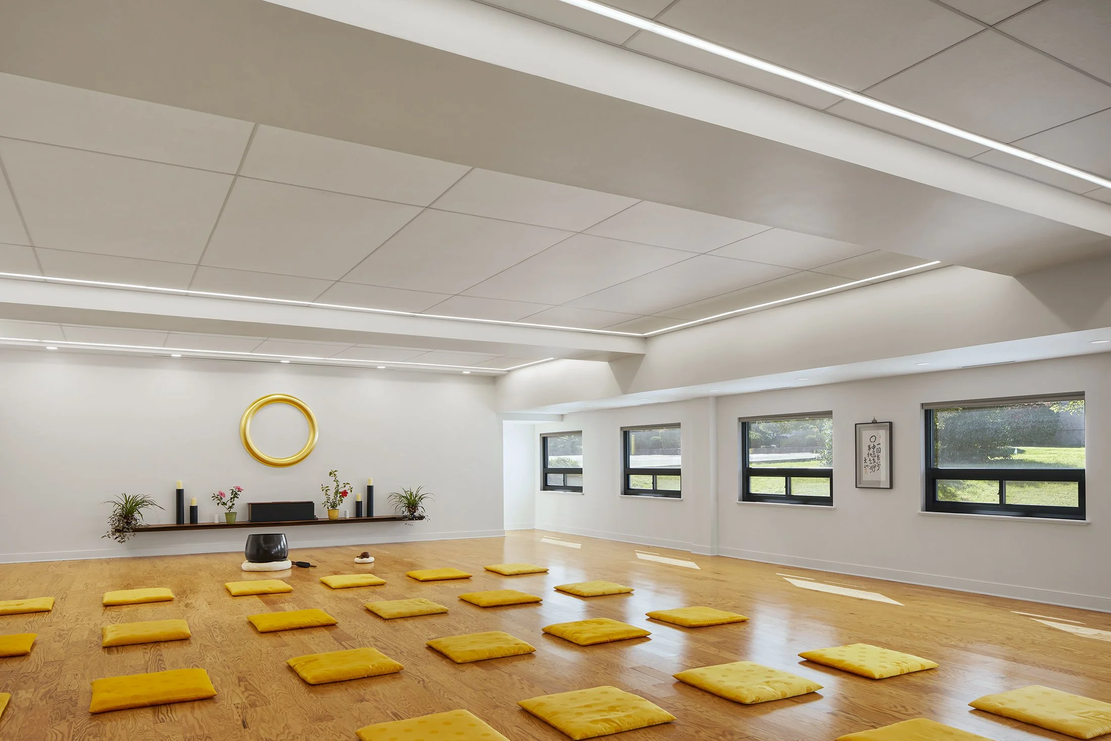 A spacious yoga or meditation studio with wooden floors, yellow cushions arranged in rows, large windows, and minimalistic decor including a wall mirror, plants, and framed artwork.