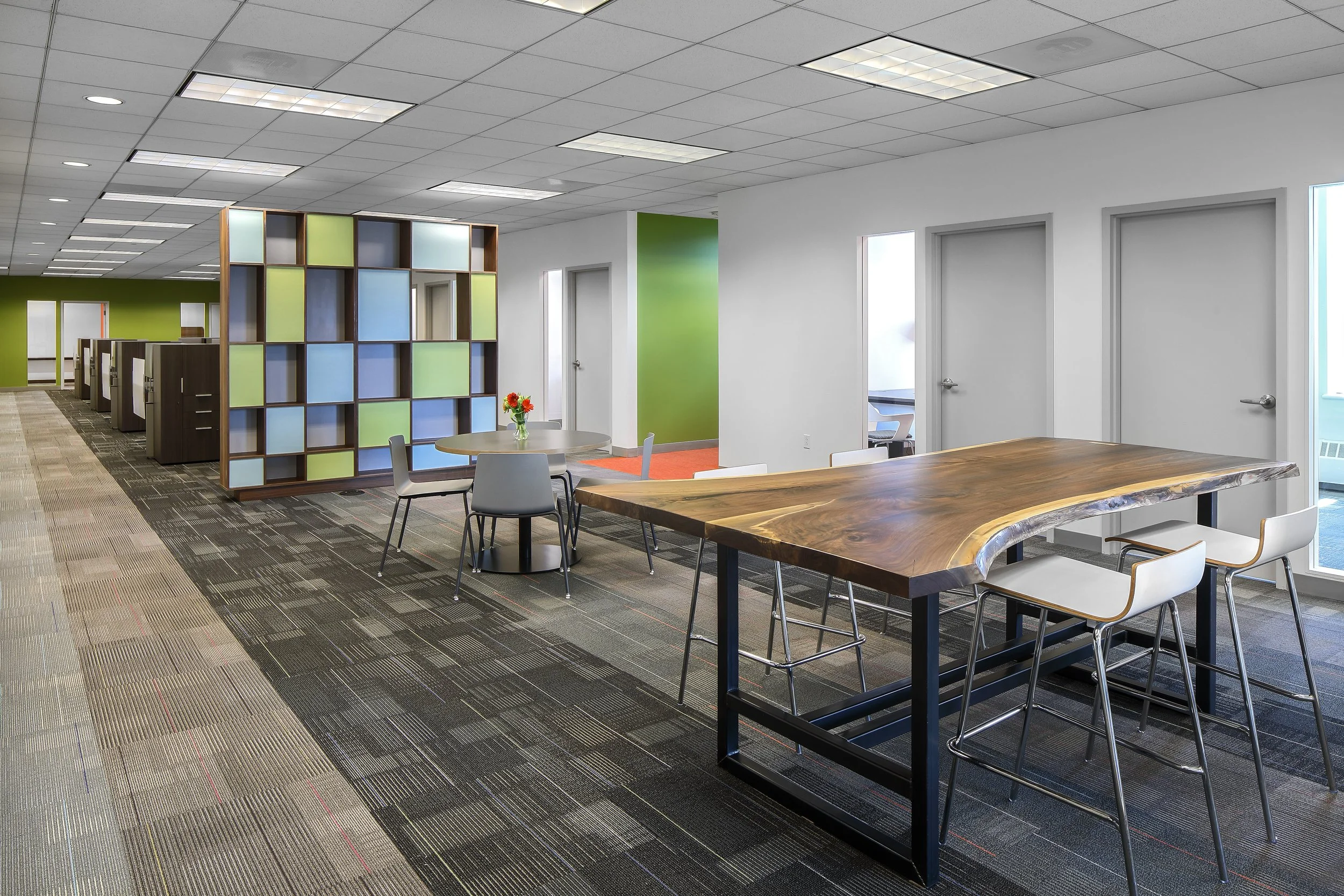 Modern office space with a large wooden table, white chairs, a round table with a flower arrangement, and partitioned workstations in the background.