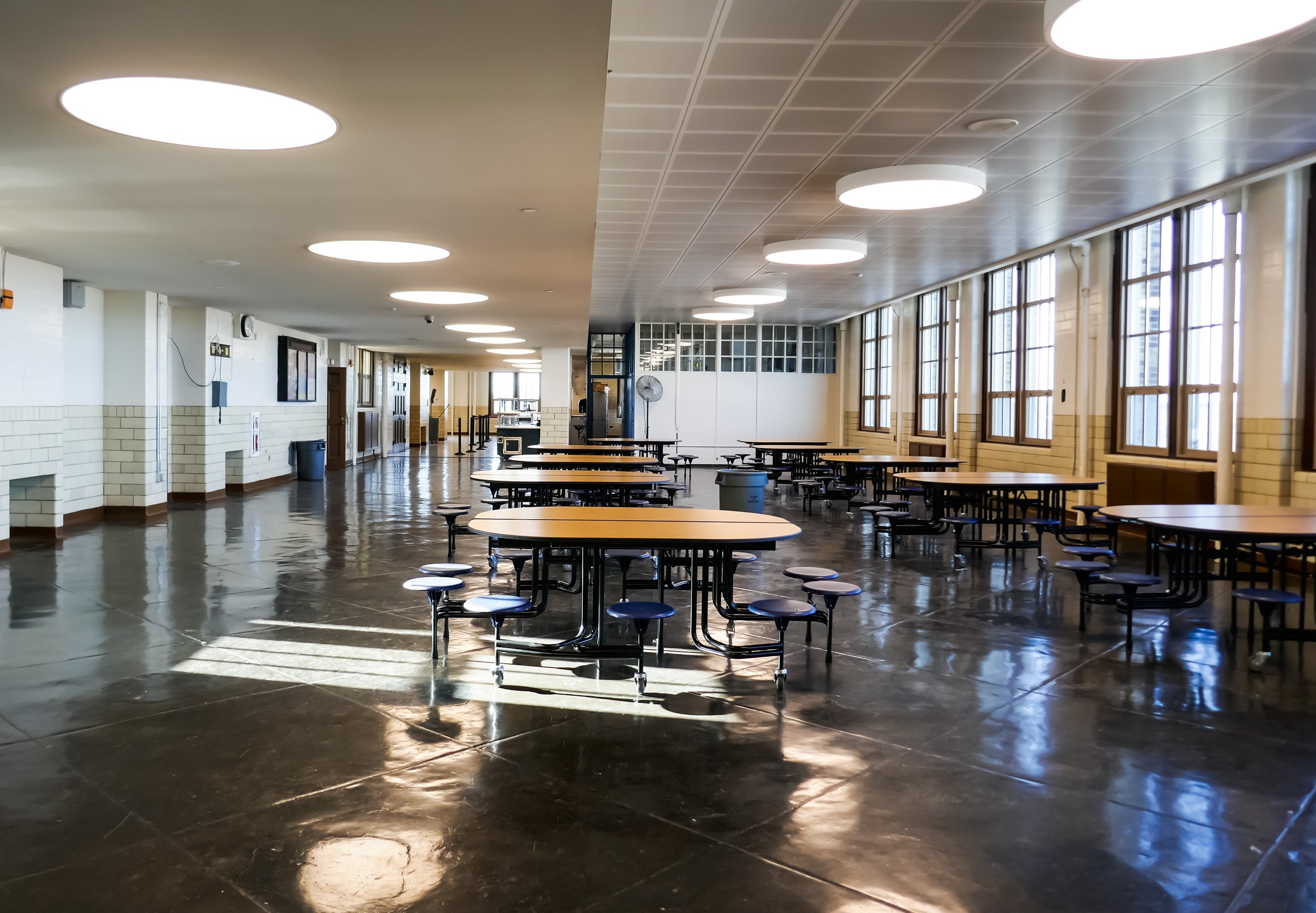 Empty school cafeteria with round tables and attached stools, large windows on the right allowing sunlight, polished dark floor, white ceiling with circular lights, walls with beige brick accents, and a few objects like a trash bin and clock in the b