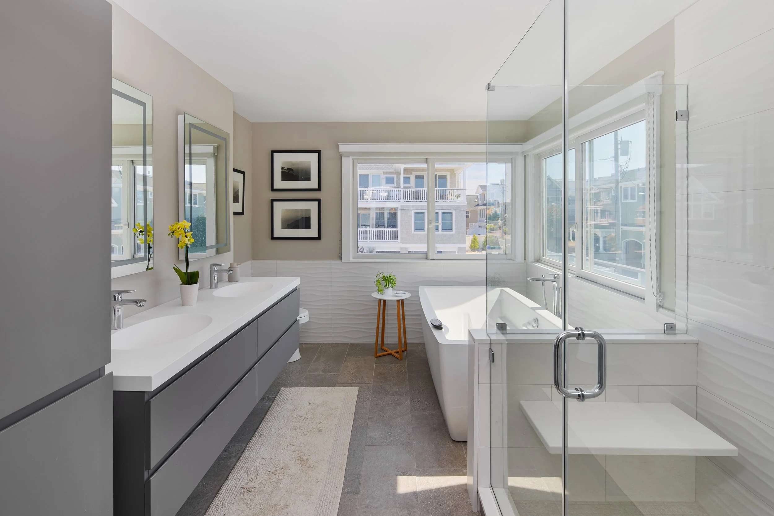 Modern bathroom with dual sinks, framed mirrors, black and white picture frames on beige wall, large windows, white bathtub, small table with a plant, beige rug, and glass shower enclosure.