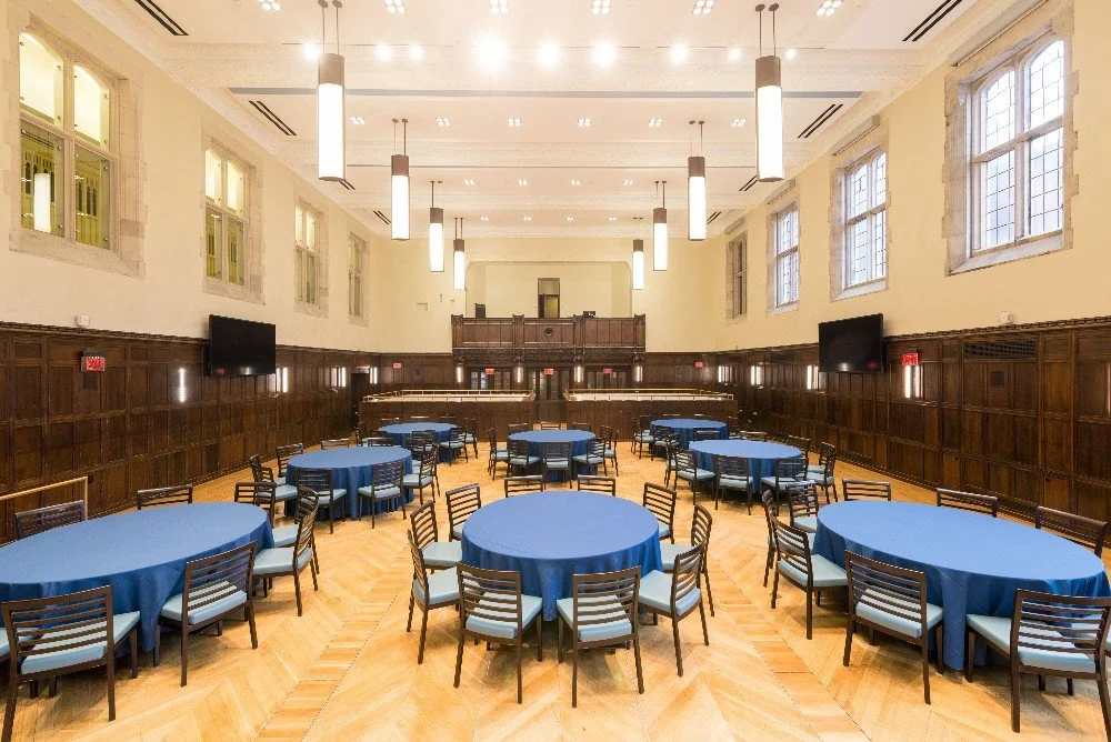 Large event hall with high ceilings, wooden panel walls, multiple round tables with blue tablecloths, and black chairs with white cushions. Windows on the upper walls and large pendant lights hanging from the ceiling.