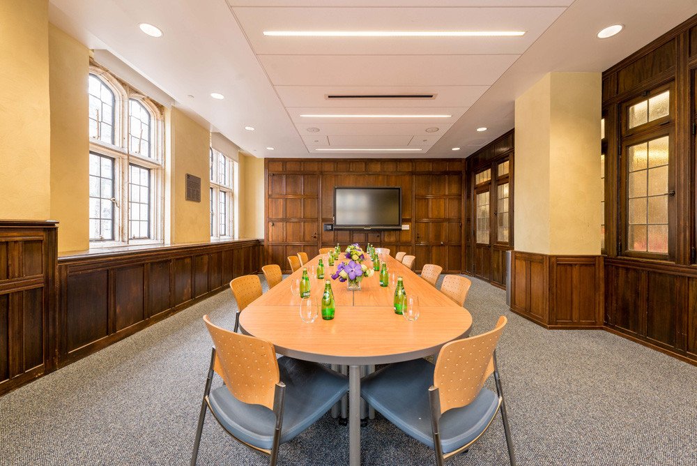 Empty conference room with a long oval table, chairs, flower arrangement, windows, and a large monitor.