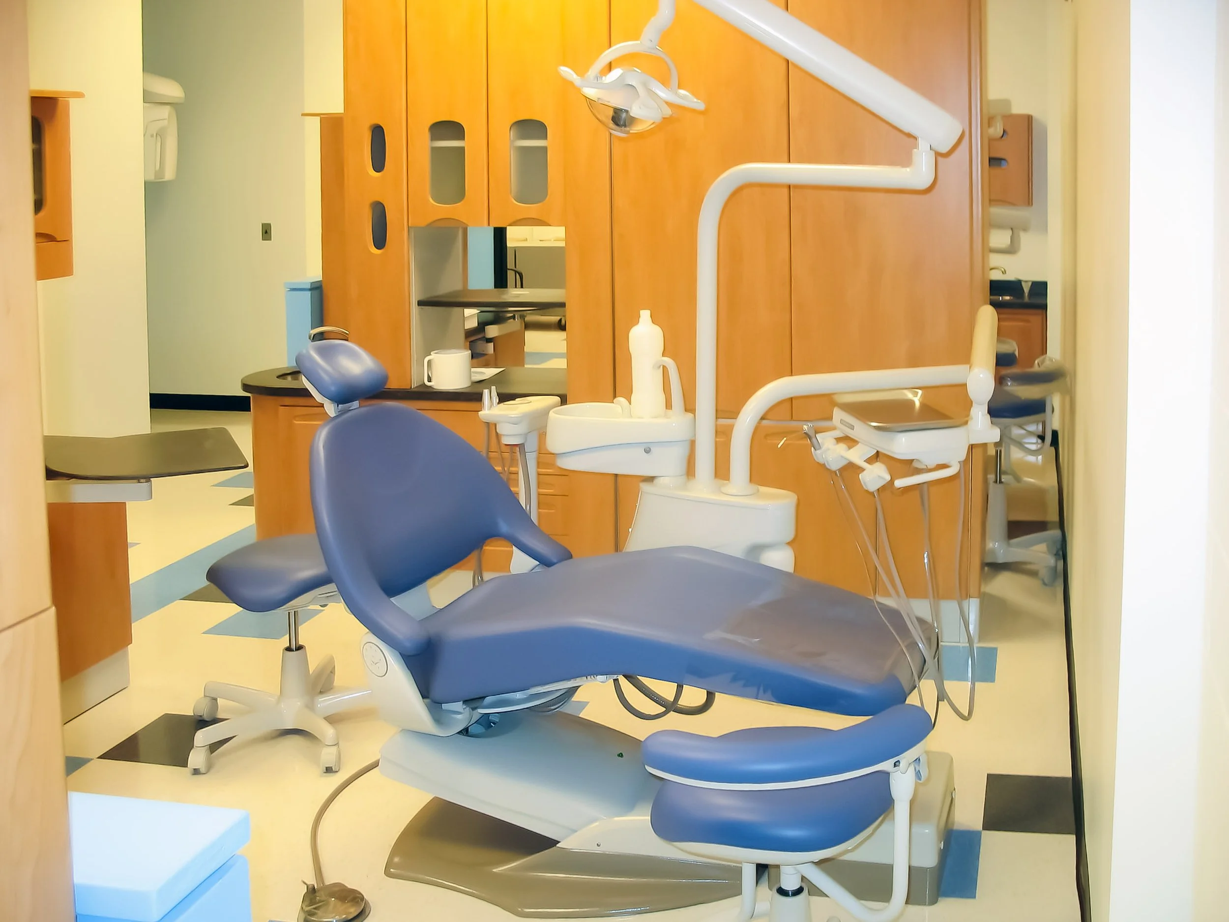 An empty dental clinic or dentist's office with a blue dental chair, various dental equipment, and cabinets in the background.