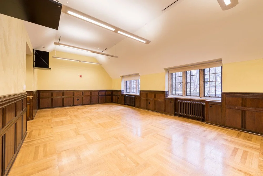 An empty room with wooden floors and paneling, yellow walls, large windows with grilles, radiators beneath the windows, and overhead fluorescent lighting.