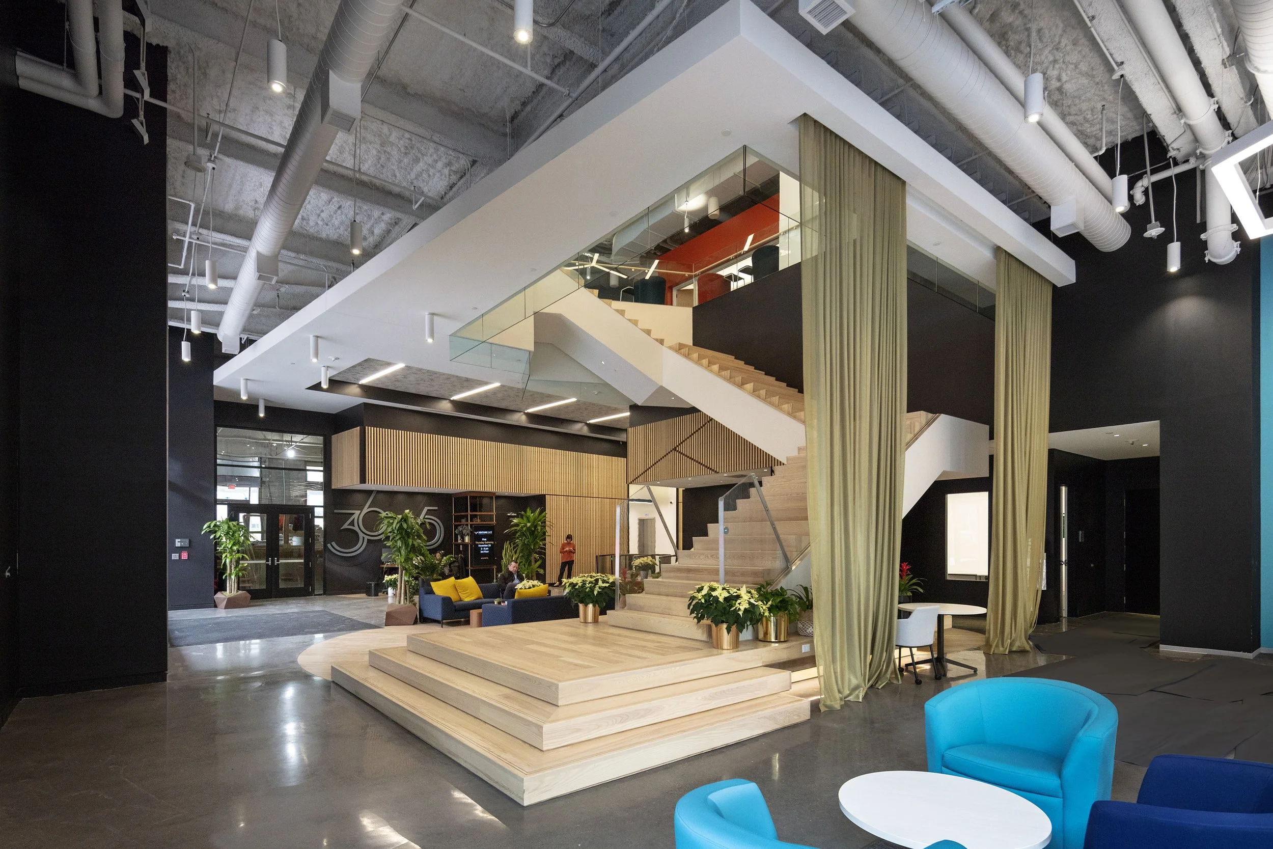 Modern hotel lobby with wooden stairs, glass railing, black walls, green curtains, blue and yellow seating areas, potted plants, and ceiling ductwork.