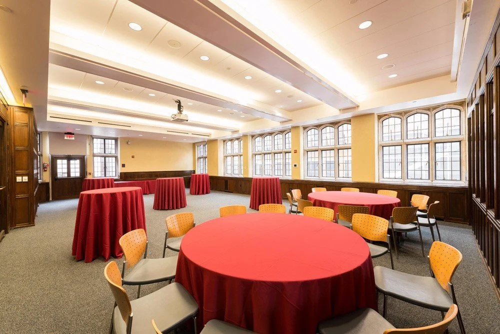 Eagerly cleaned banquet hall with round tables covered in red tablecloths and matching high-top tables, accented with beige chairs, large windows, warm lighting, and wooden wall paneling.
