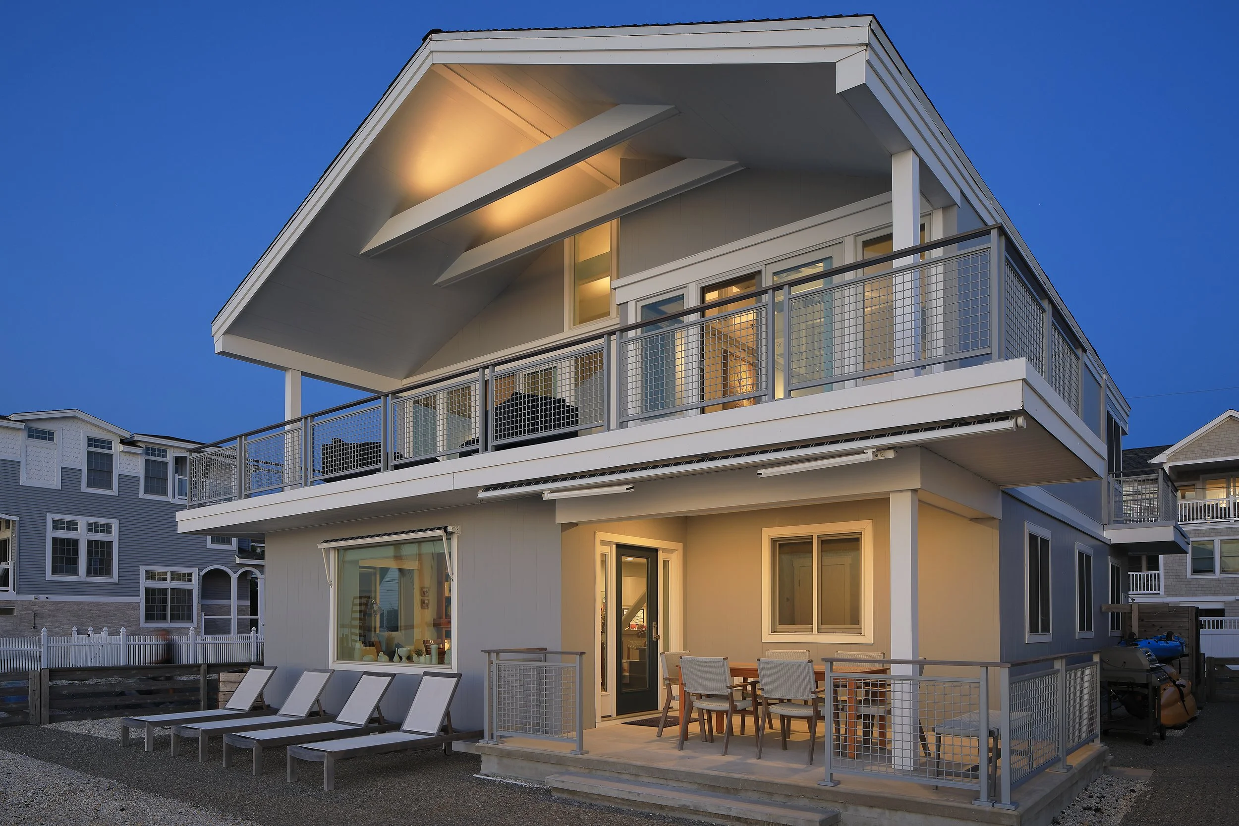 Two-story modern house with large balcony on the upper floor and patio on the ground level, illuminated at dusk, with outdoor seating and lounge chairs.