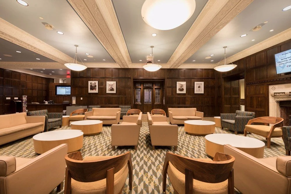 A spacious hotel lobby or lounge with beige and patterned furniture, wooden wall paneling, framed black-and-white portraits, and a fireplace on the right.