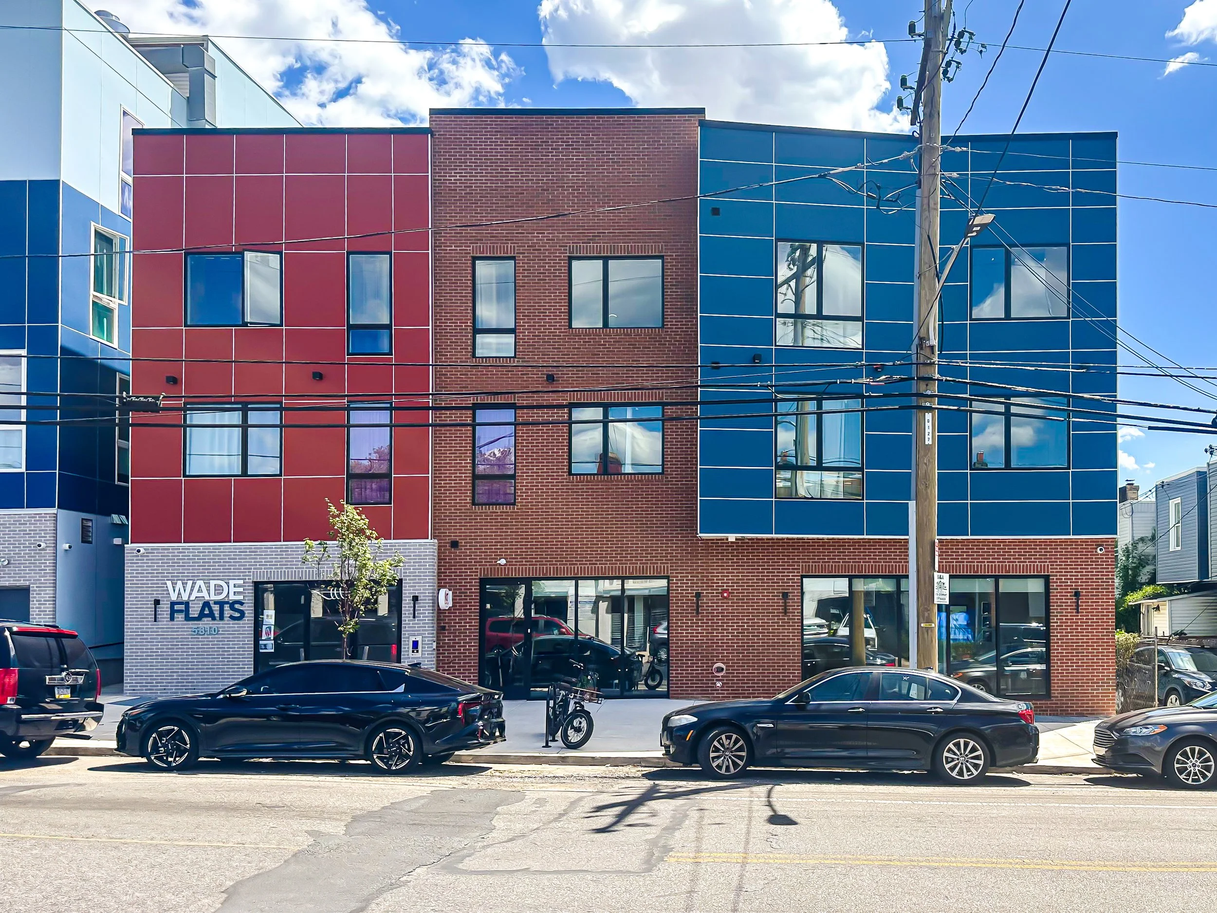 A modern red, blue, and gray apartment building with large windows, parking along the street, and a power pole in front on a sunny day with partly cloudy sky.